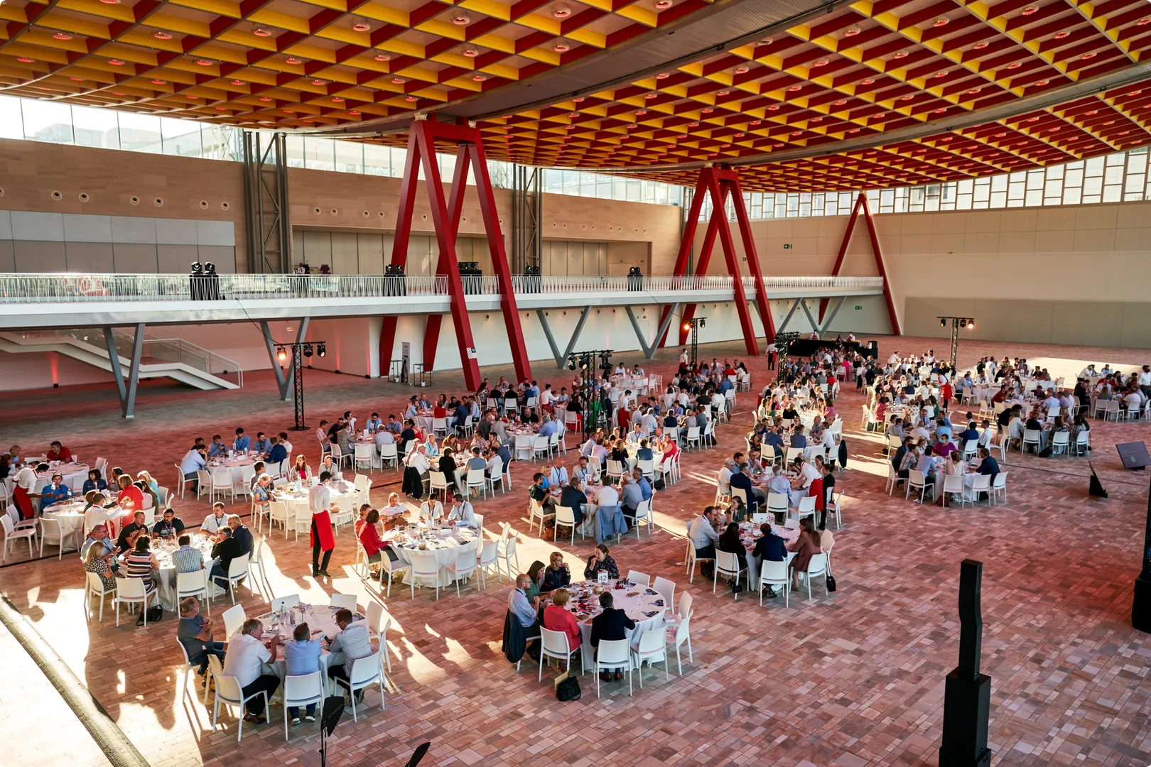 Large indoor venue with red beams and a checkered ceiling, filled with round tables and people seated, engaging in conversation.