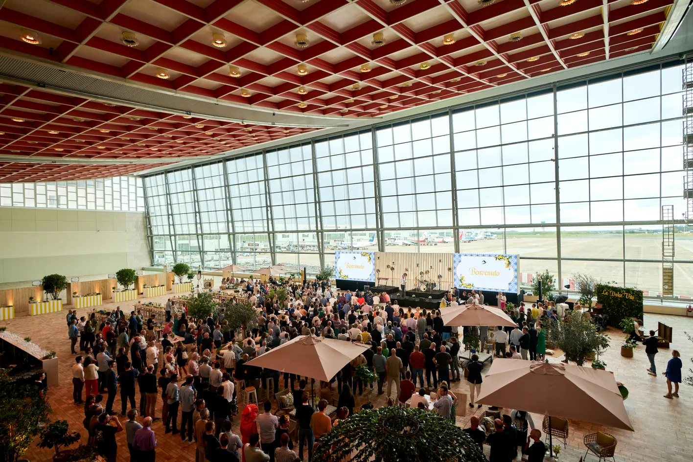 A large hall with glass walls is filled with a crowd facing a stage, decorated with plants and umbrellas, and a "Benvenuto" sign is displayed.