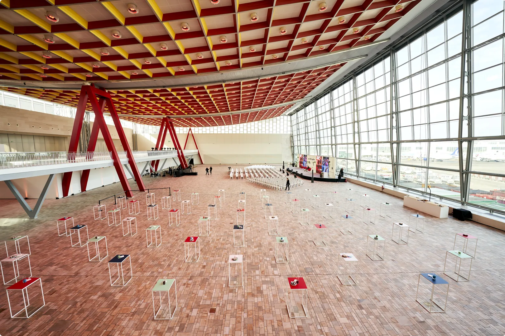 Spacious modern event hall with a geometric red and yellow ceiling, large windows, scattered podiums, and a seating area.