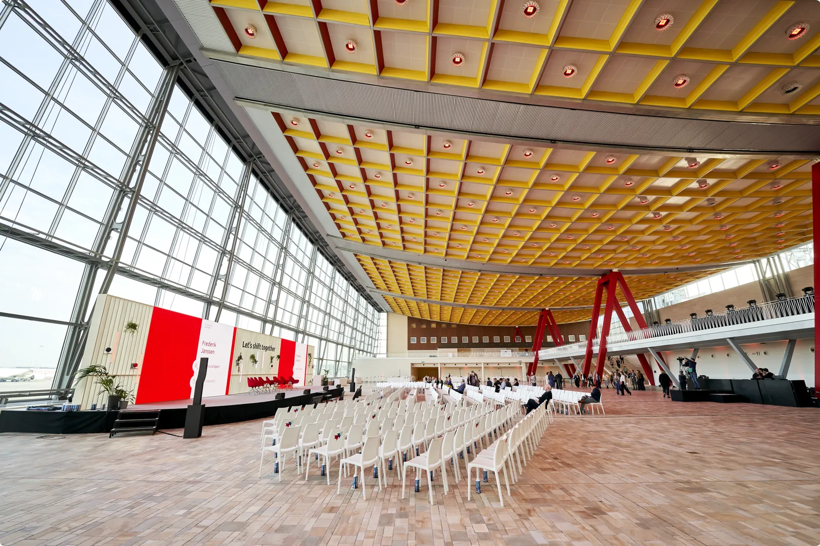 Spacious modern conference hall with colorful ceiling, large glass windows, red stage backdrop, and rows of white chairs arranged for an event.