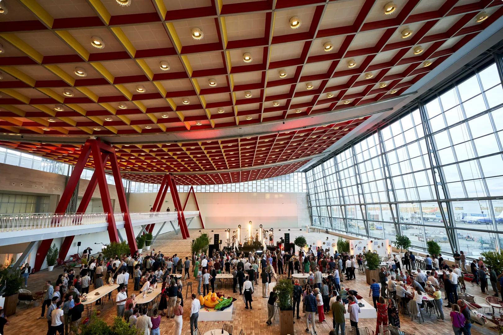 A crowded modern event space with high ceilings, large windows, red structural beams, and people mingling around tables.