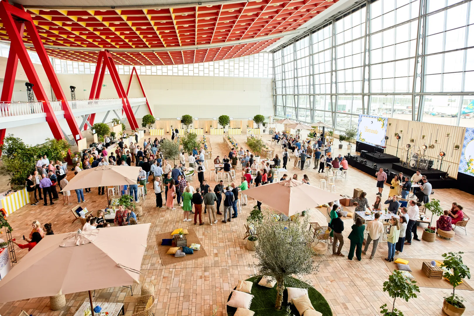 A large indoor event with people mingling under umbrellas, surrounded by plants, with a bright, high-ceilinged atrium.