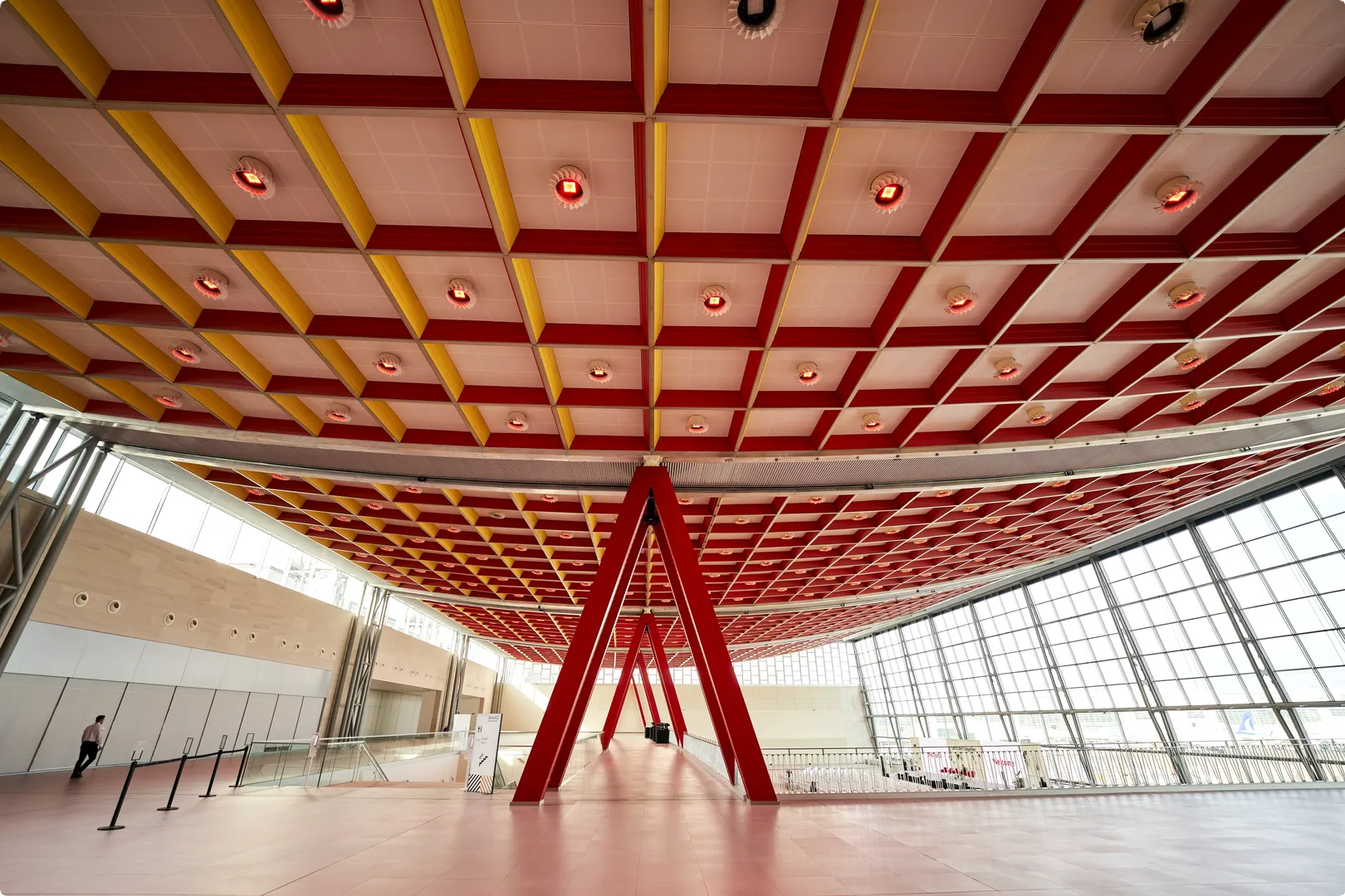 Spacious modern interior with a red and yellow geometric ceiling, a large windowed wall, and a central triangular support structure.