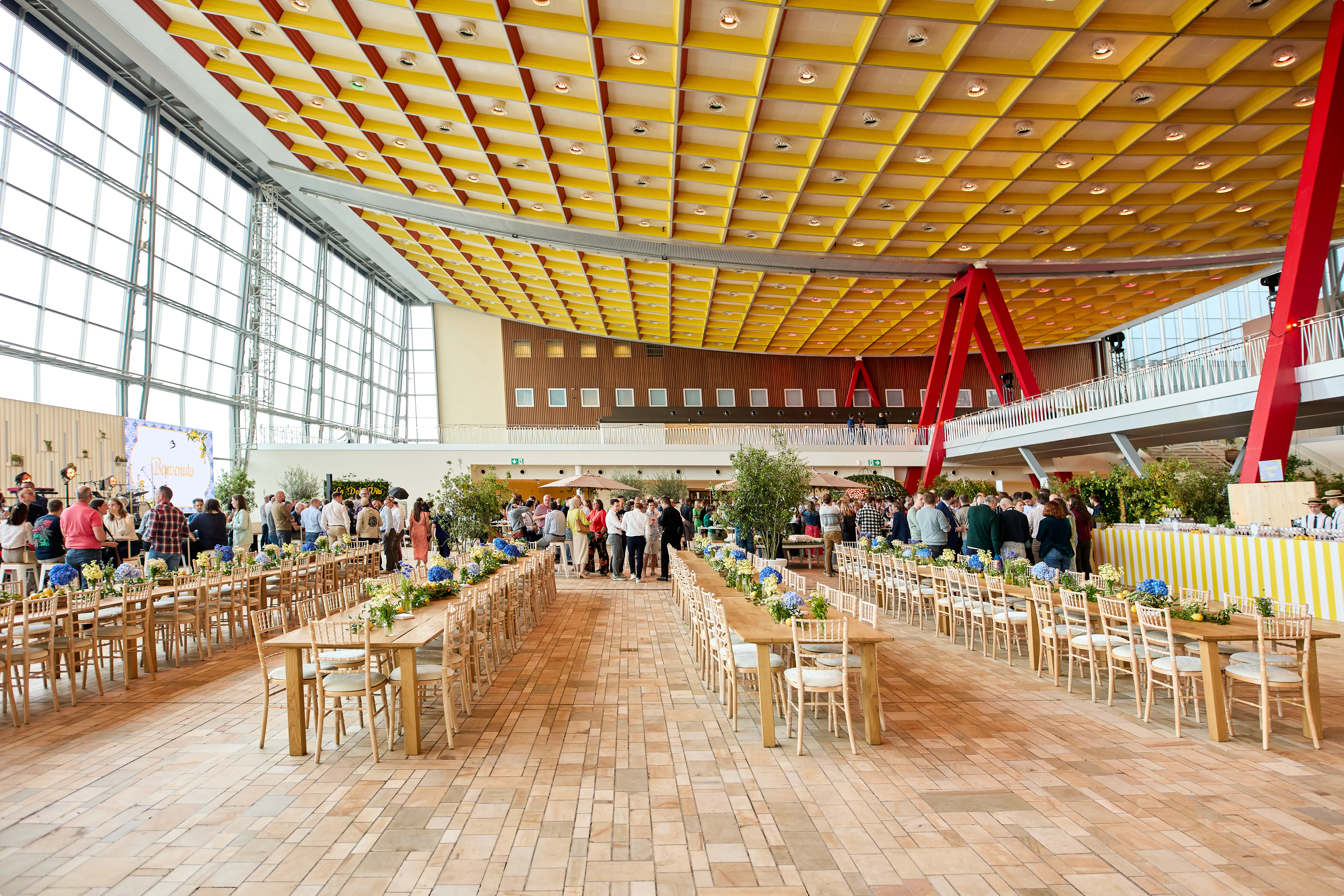 Spacious event hall with colorful ceiling, wooden tables set for dining, and a crowd of people gathered near the entrance.