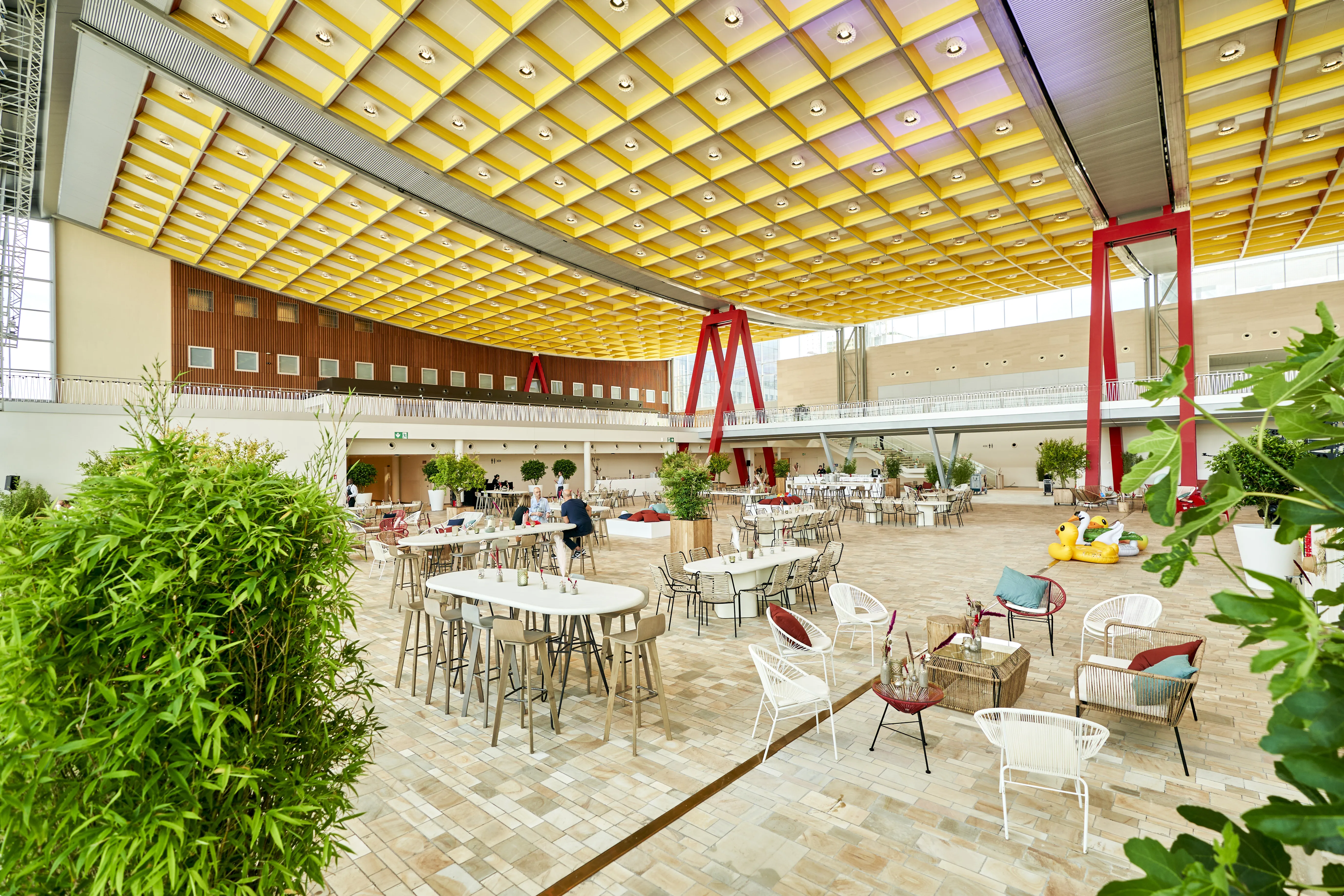 Spacious indoor atrium with yellow grid ceiling, red columns, tables and chairs, lush plants, and inflatable pool toys.