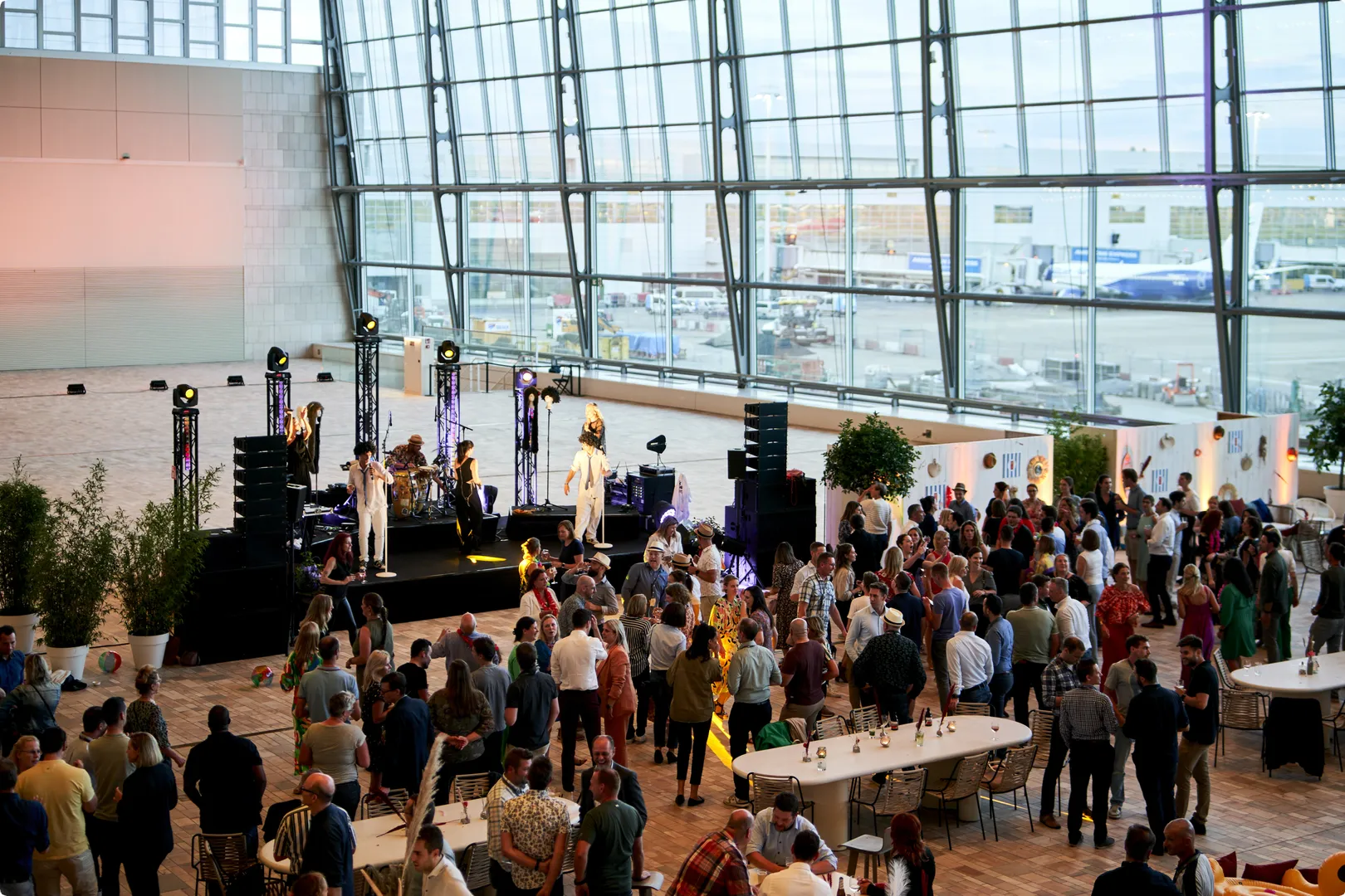 A crowd gathers in a modern, spacious venue with large glass windows, a stage with performers, and tables set up around the room.