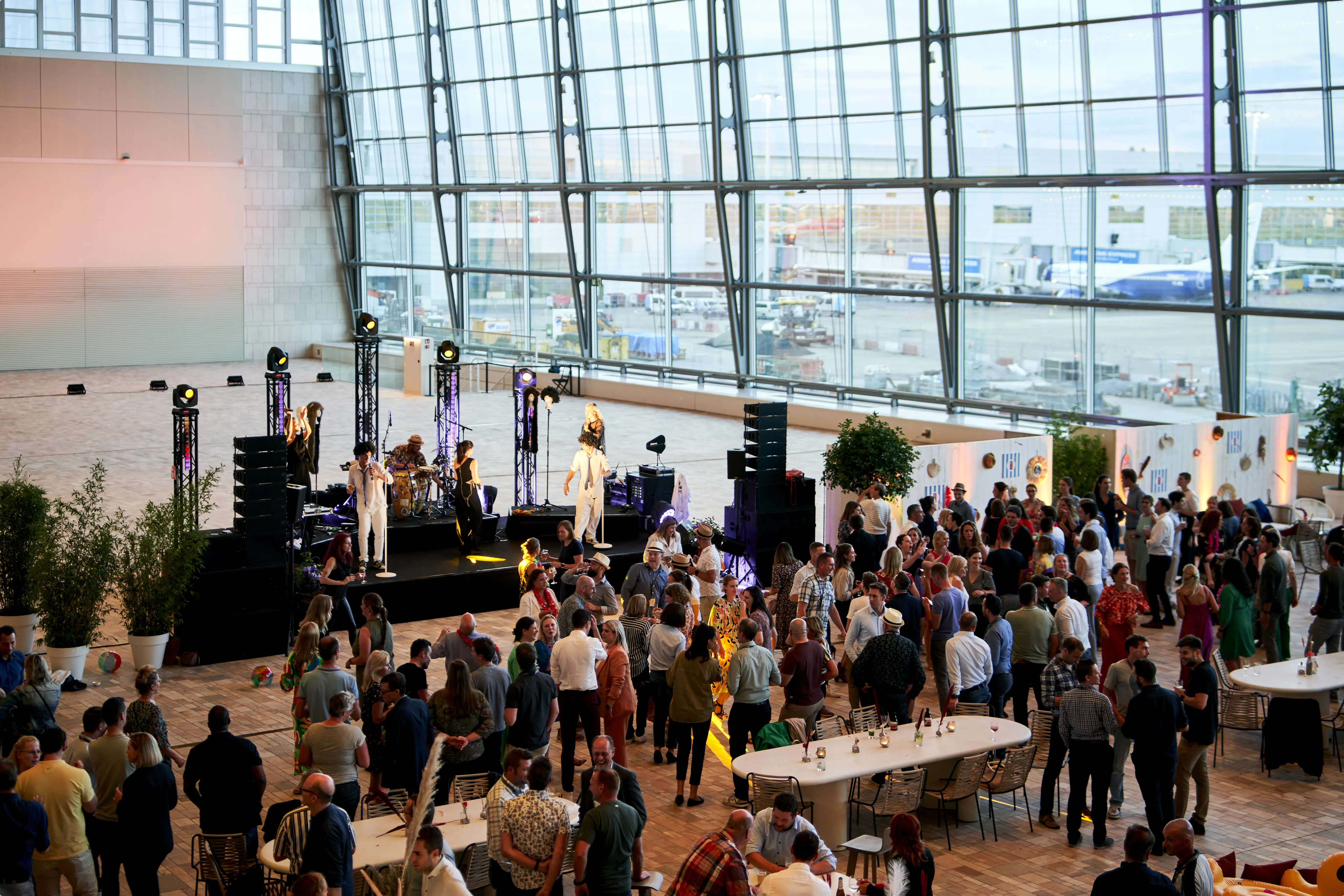 A crowd gathers in a modern, spacious venue with large glass windows, a stage with performers, and tables set up around the room.