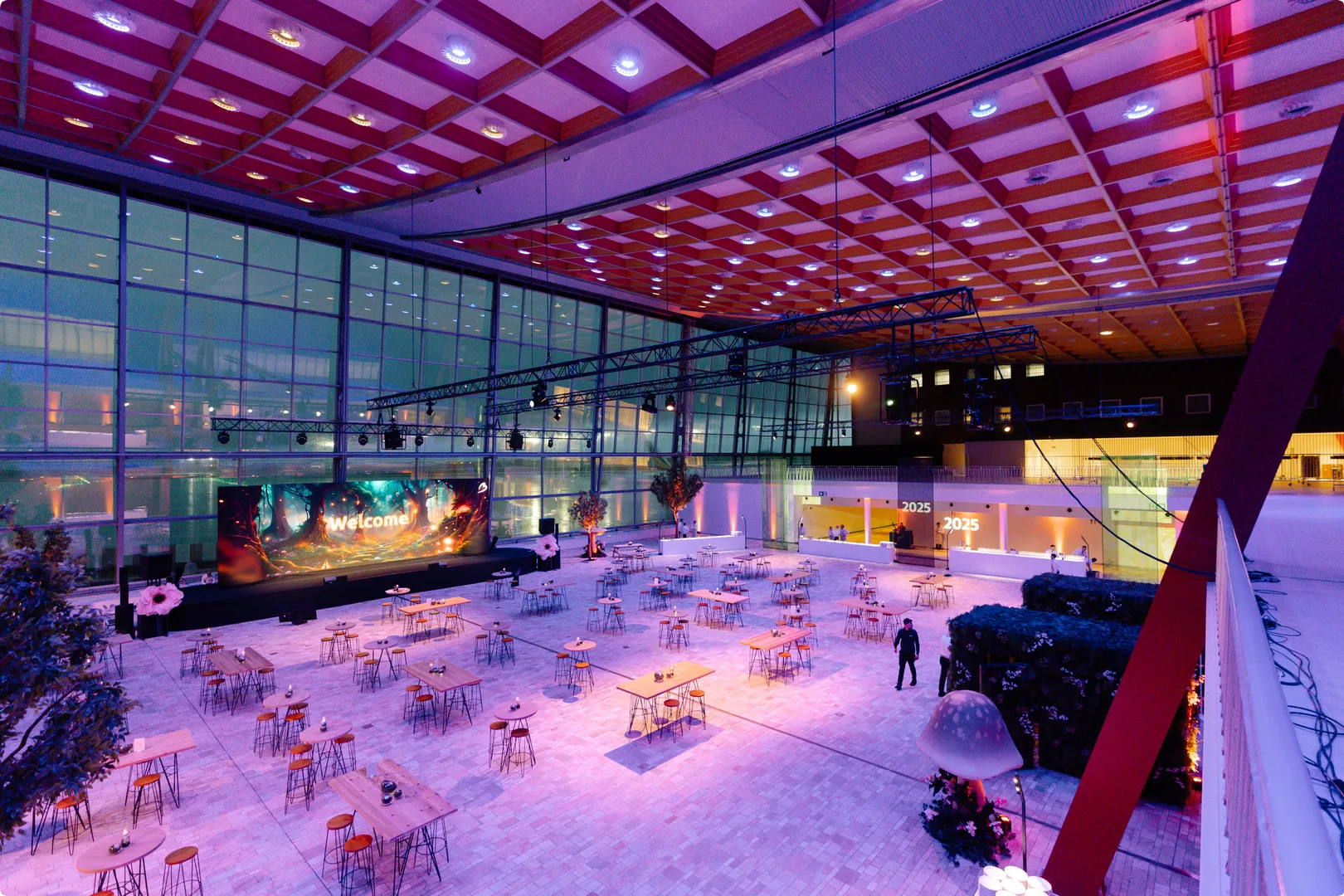 Spacious, modern hall with glass walls and ceiling featuring a large "Welcome!" sign, illuminated tables, and decorative plants.
