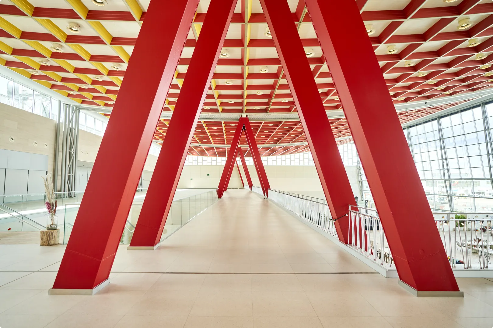 Interior of a modern building with large red angled beams and a patterned ceiling, leading to a bright windowed area.