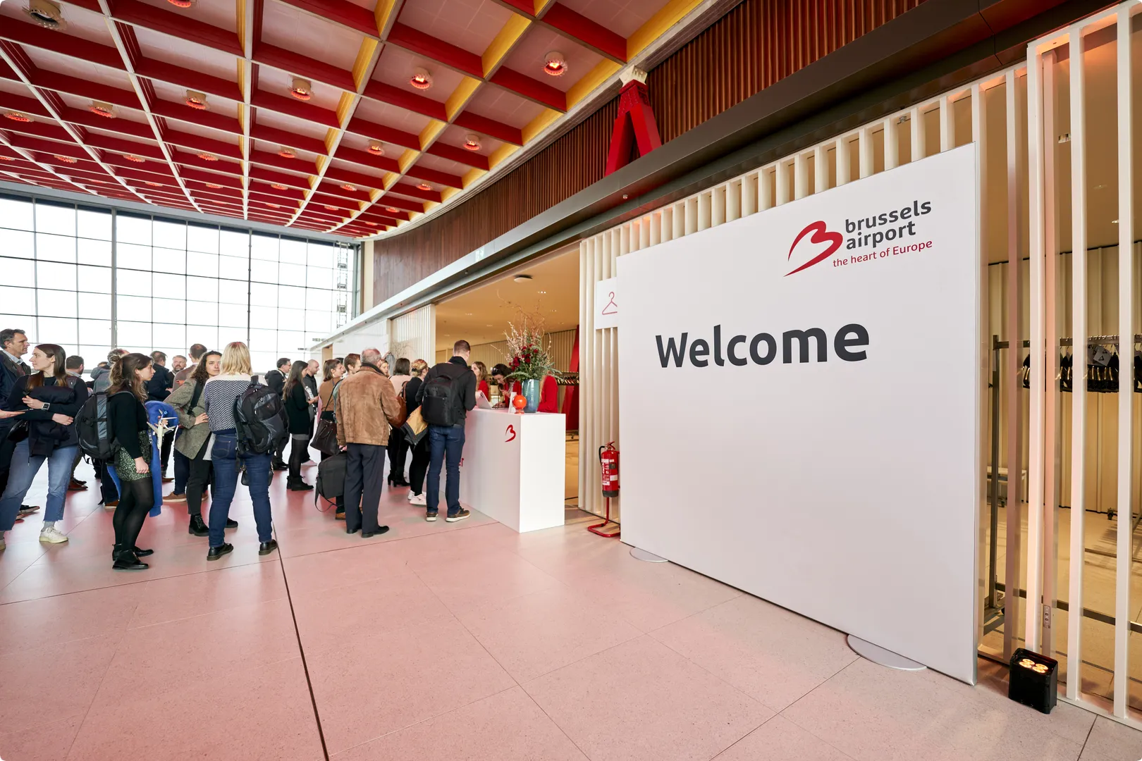 Crowd gathered near a welcome sign at Brussels Airport, with a modern interior and a vibrant red and yellow ceiling pattern.