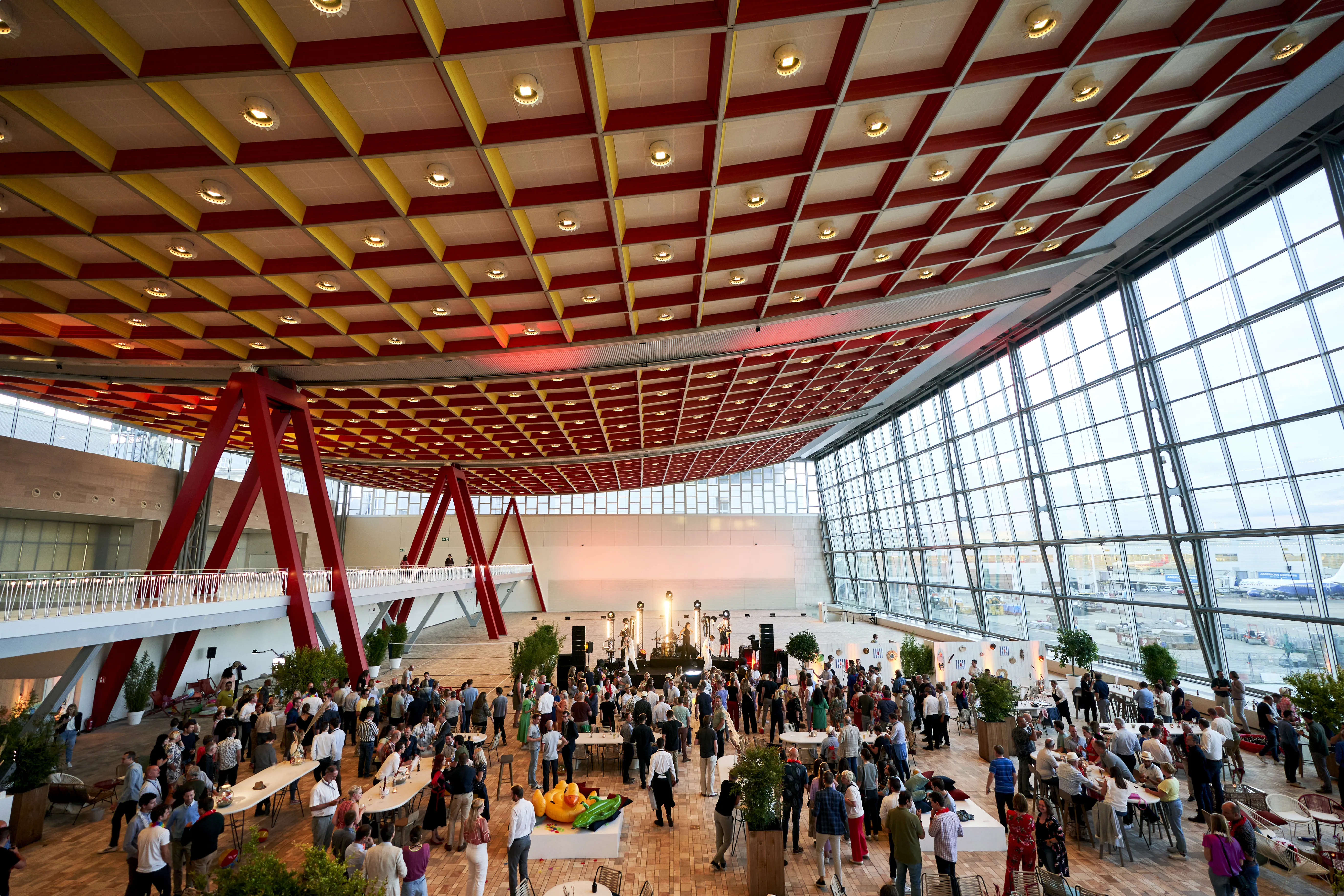 A spacious indoor venue with a colorful ceiling, large windows, and a crowd attending an event with tables, greenery, and a stage under bright lights.