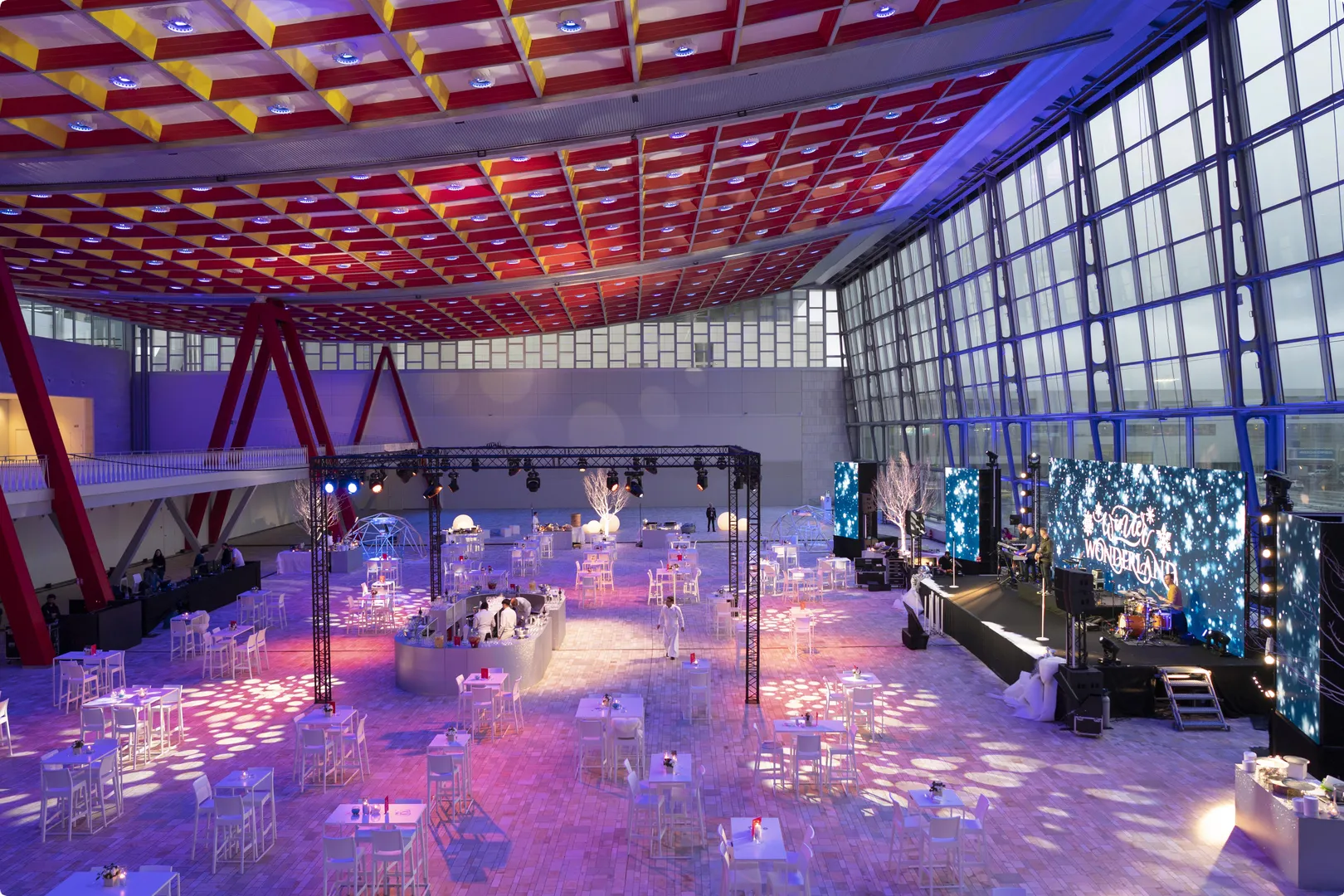Spacious indoor event venue with colorful ceiling, dining tables, a stage with "Winter Wonderland" display, and ambient lighting.