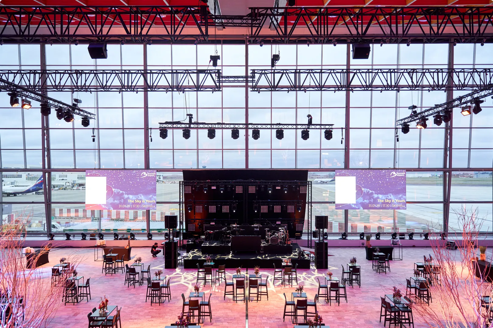 Spacious event hall with stage setup, lighting rigs, tables, and chairs, overlooking an airport runway through large glass windows.