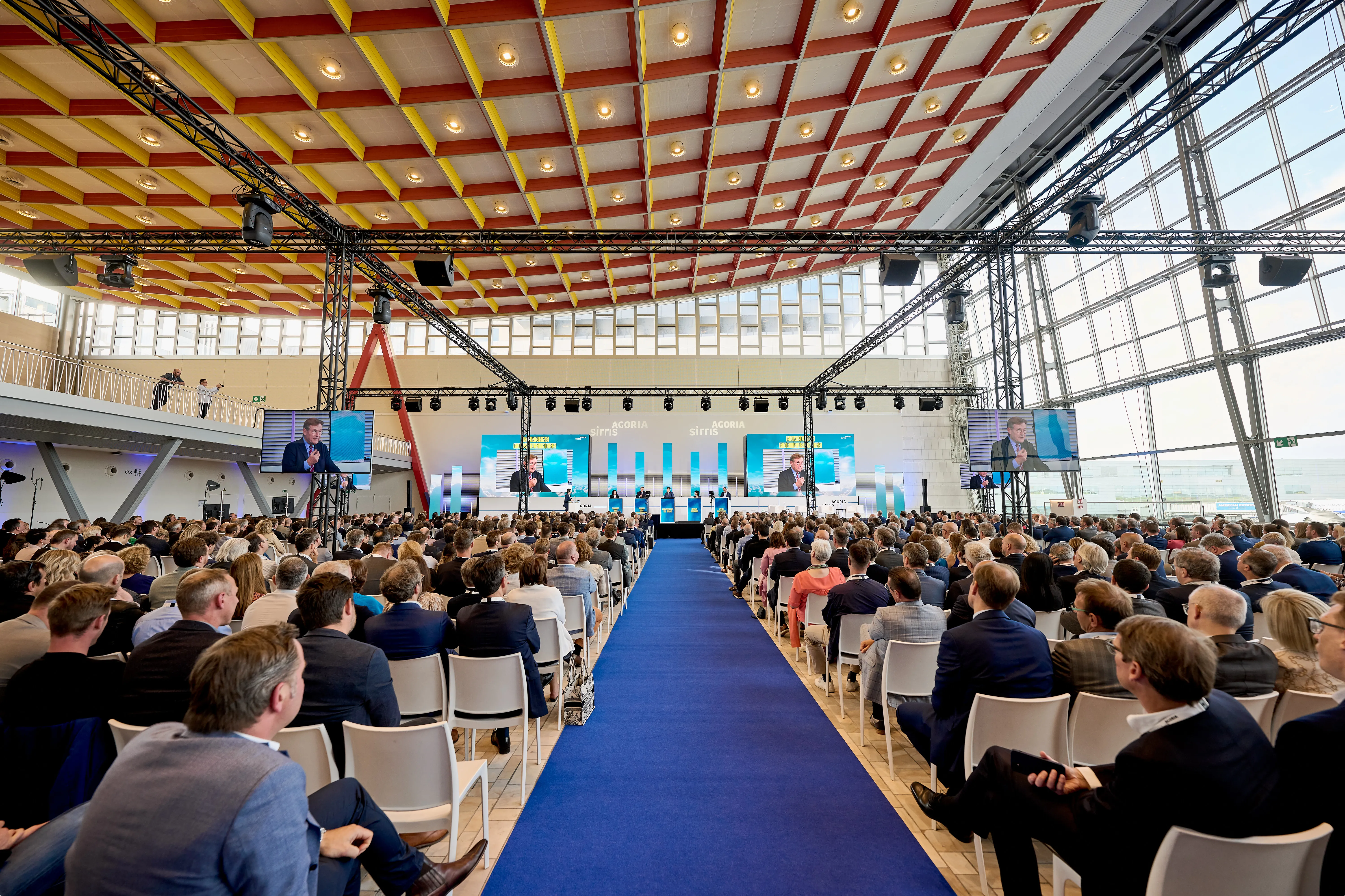 A large audience seated indoors faces a stage with multiple screens and speakers, in a spacious venue with a high ceiling and large windows.