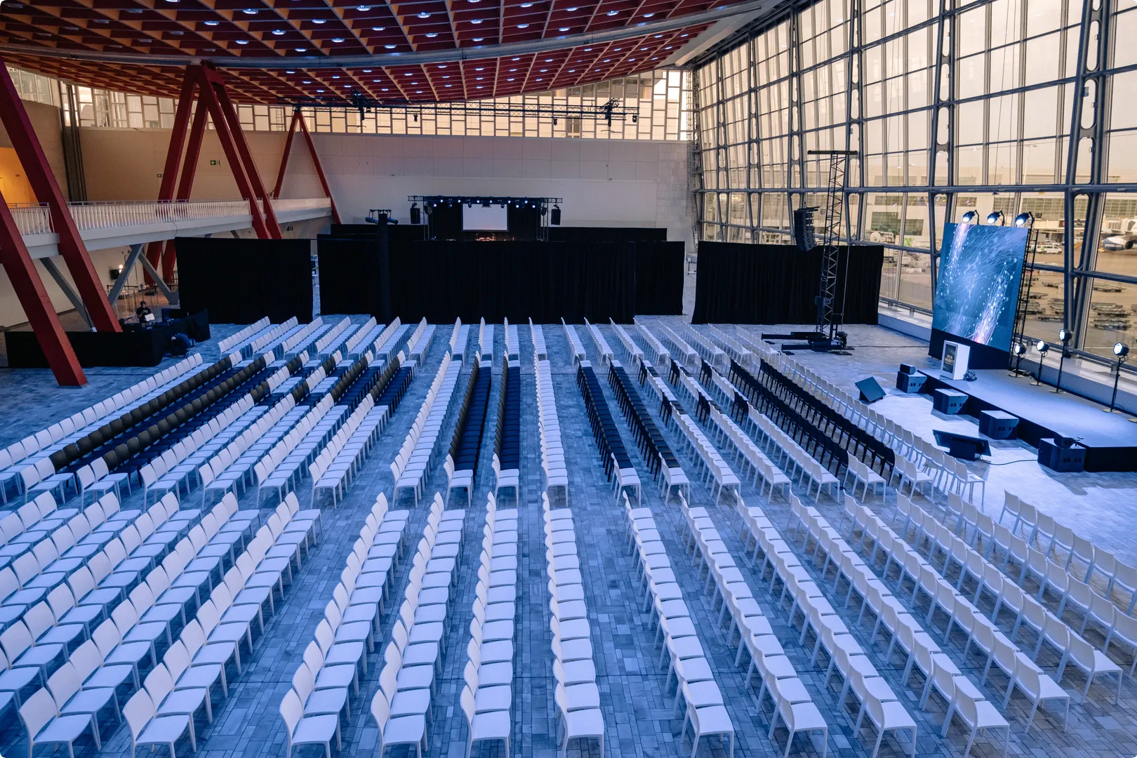 Rows of empty white chairs in a large conference hall with a stage and electronic display, surrounded by glass walls and red beams.