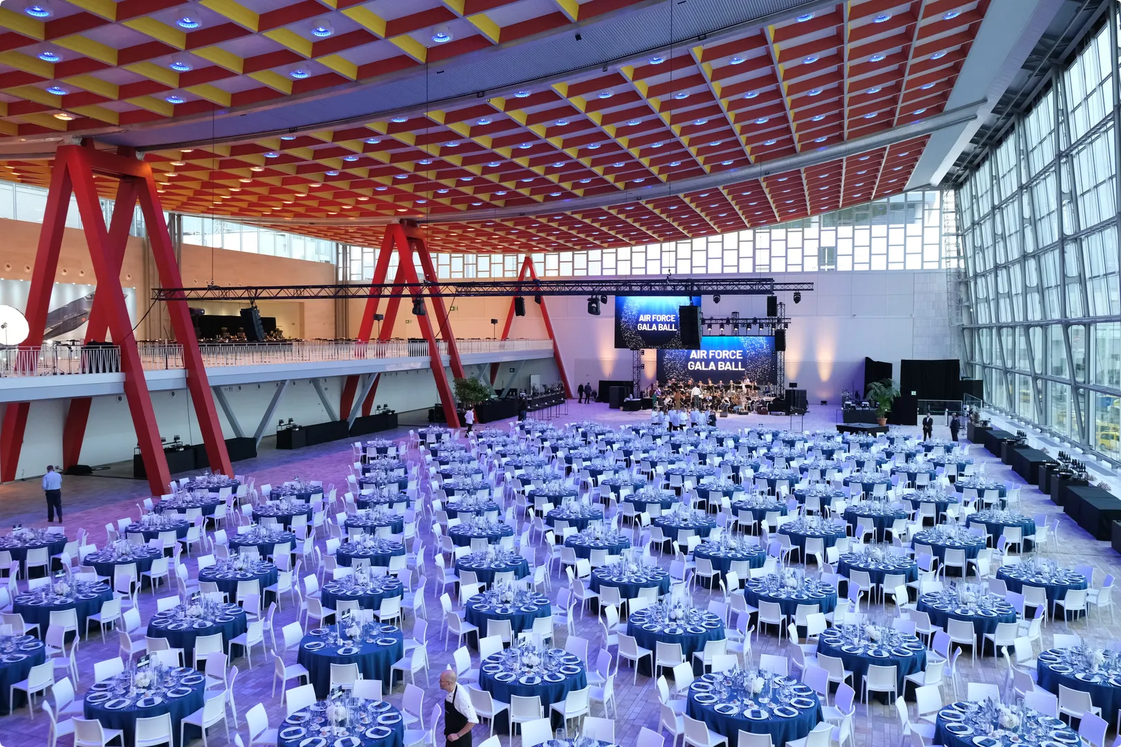 Spacious hall set for Air Force Gala Ball with numerous round tables, elegant settings, a stage, red beams, and a glass wall with a grid ceiling above.