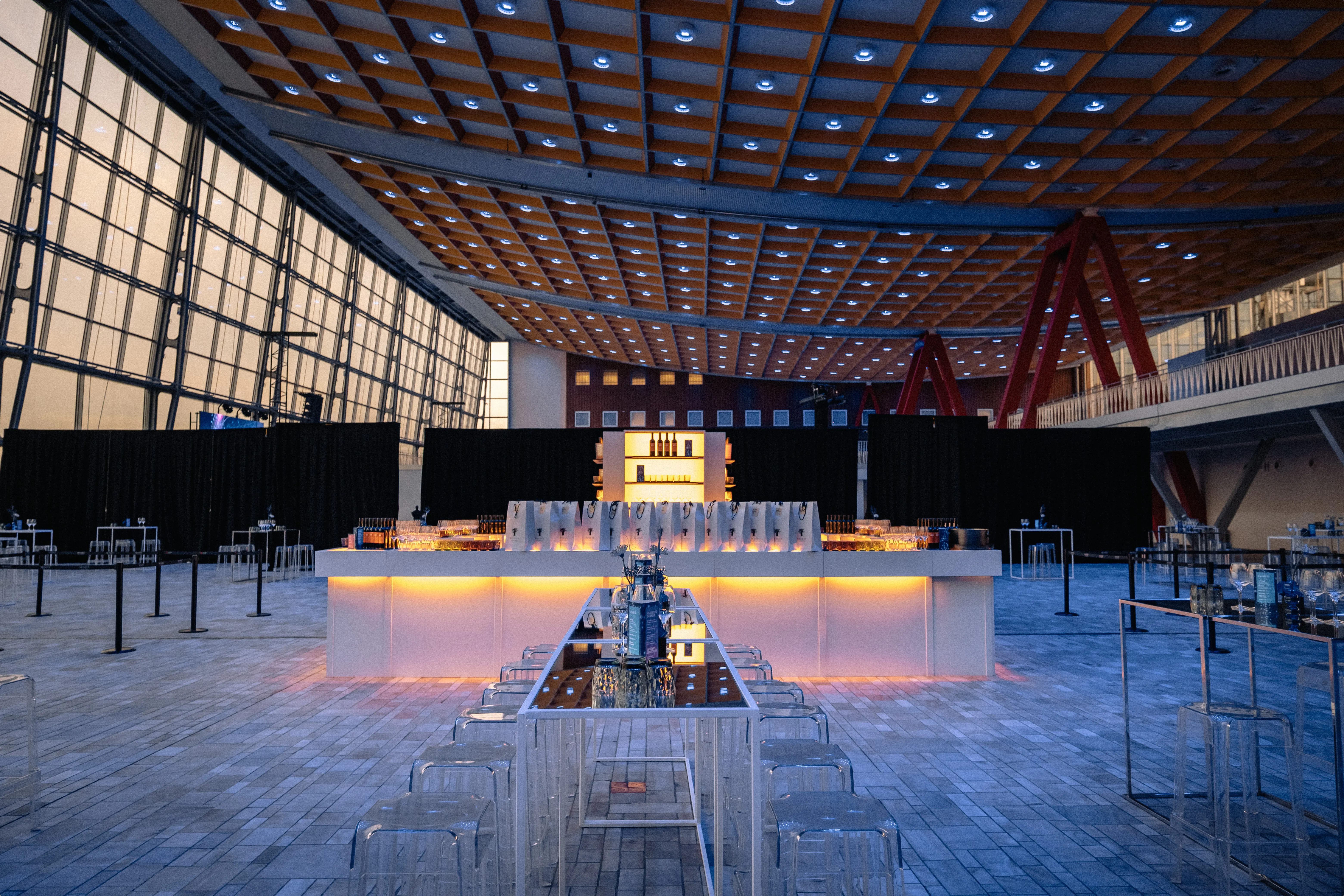 Spacious modern event hall with orange-lit bar, glass stools, and high ceiling, featuring large windows and a patterned tile floor.
