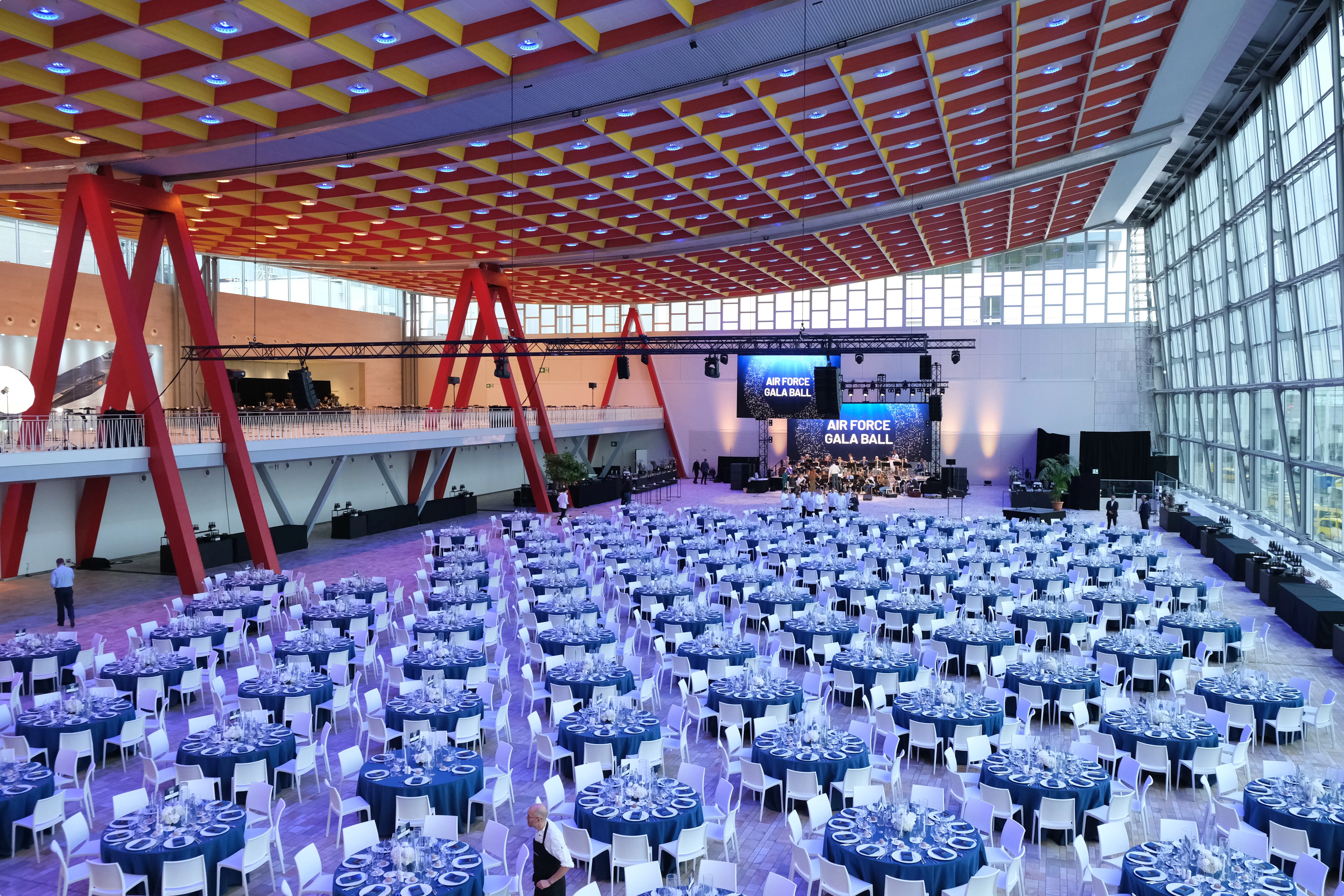 Spacious event hall with blue-lit round tables and white chairs, set up for the Air Force Gala Ball. A stage and large screens at the front.