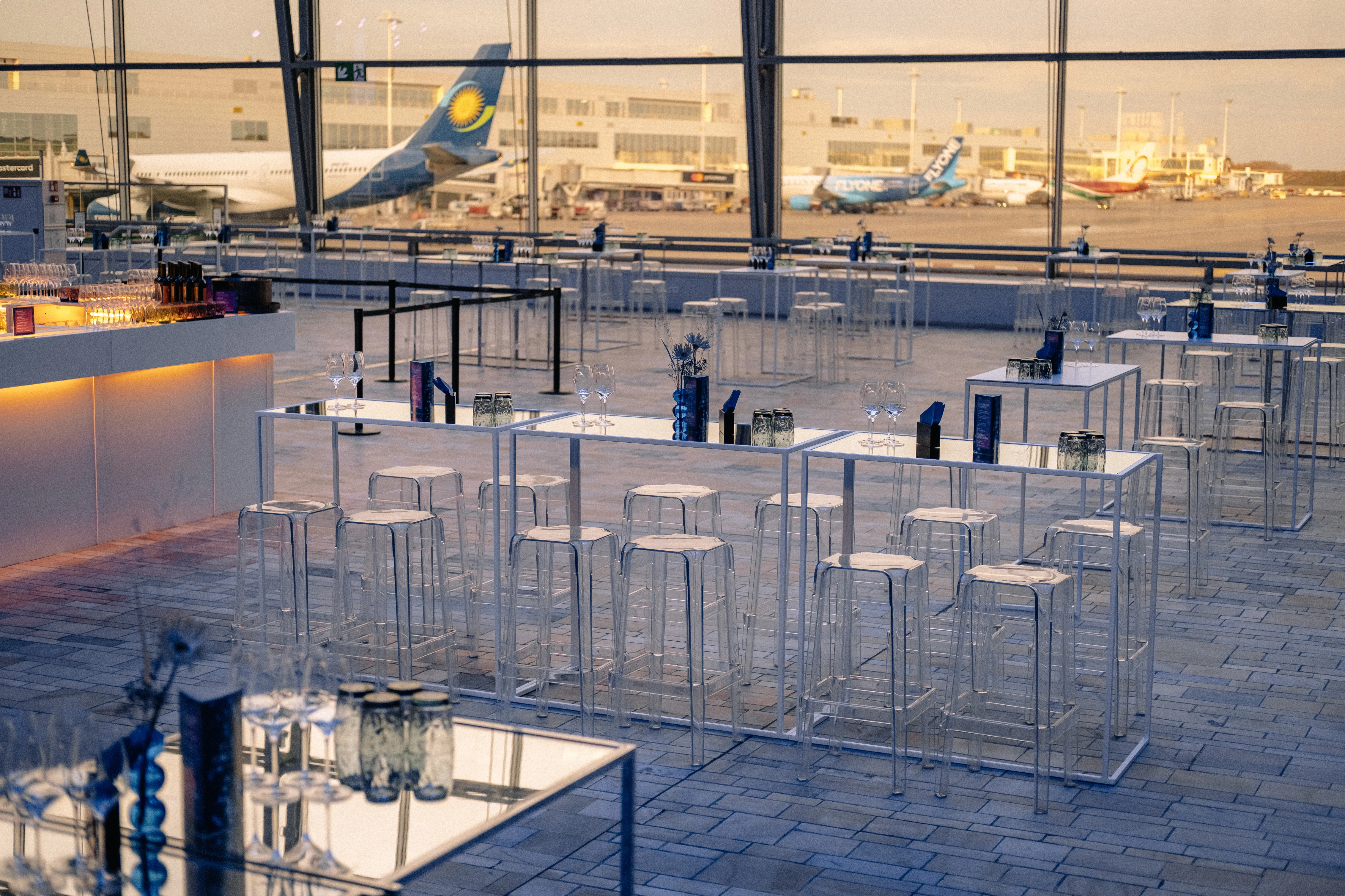 Modern airport lounge with clear tables and stools, ambient lighting, and aircraft visible through large windows in the background.