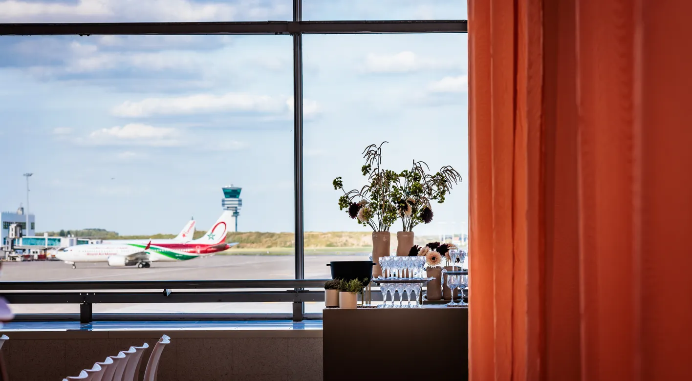 View of an airport runway through a large window, with a plane and control tower outside, and flowers and wine glasses on a table indoors.