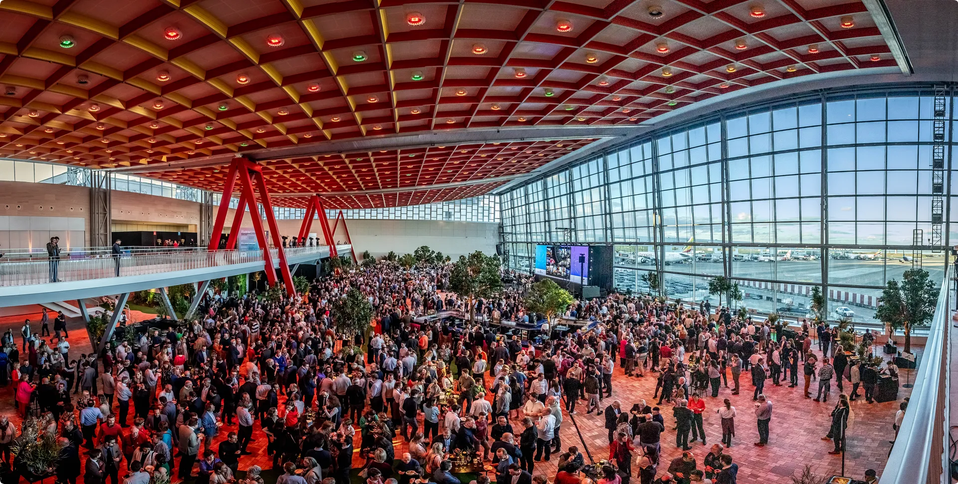 A large, bustling crowd gathers in a modern, spacious airport terminal with a high ceiling and expansive windows overlooking the tarmac.