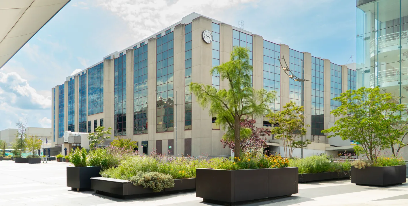 Modern building with mirrored windows, surrounding planters with lush greenery, and a bright blue sky with scattered clouds above.