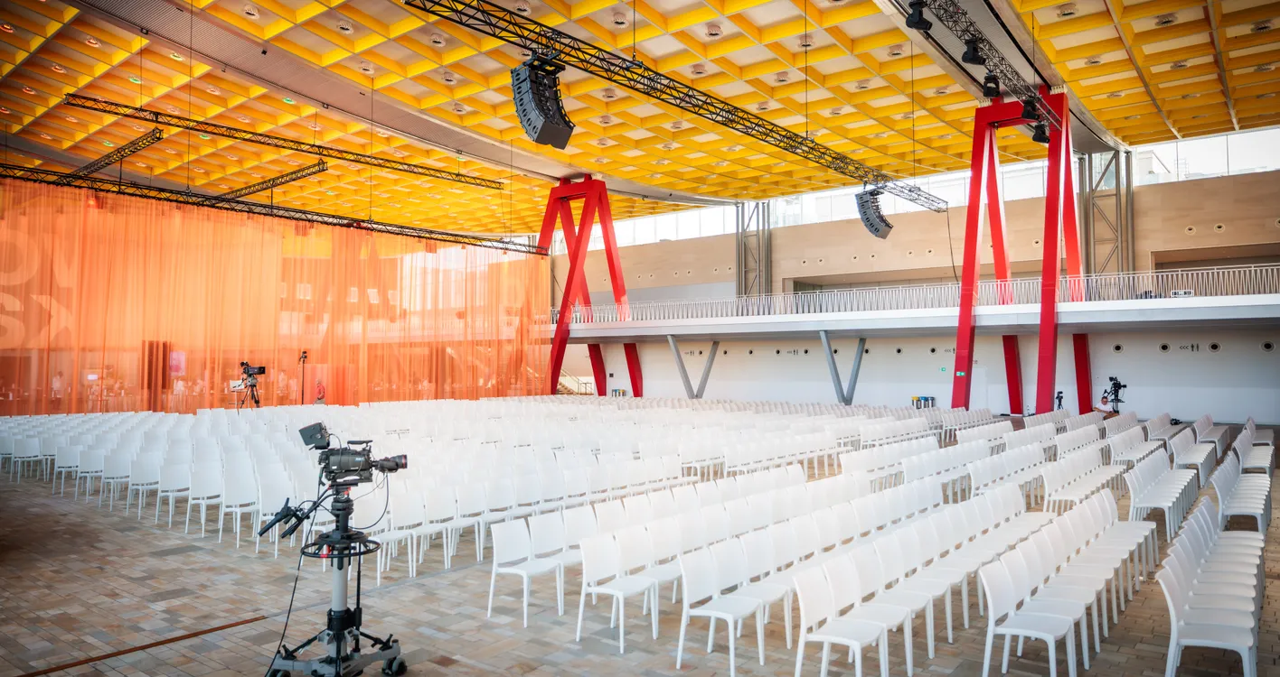 Empty modern conference hall with white chairs, orange drapes, yellow ceiling, red support beams, and cameras set up for filming.