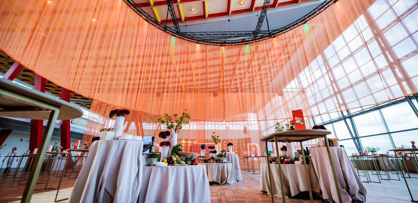 A modern interior with orange sheer drapes hanging from the ceiling, tables with white cloths, flowers, and fruits displayed as decoration.