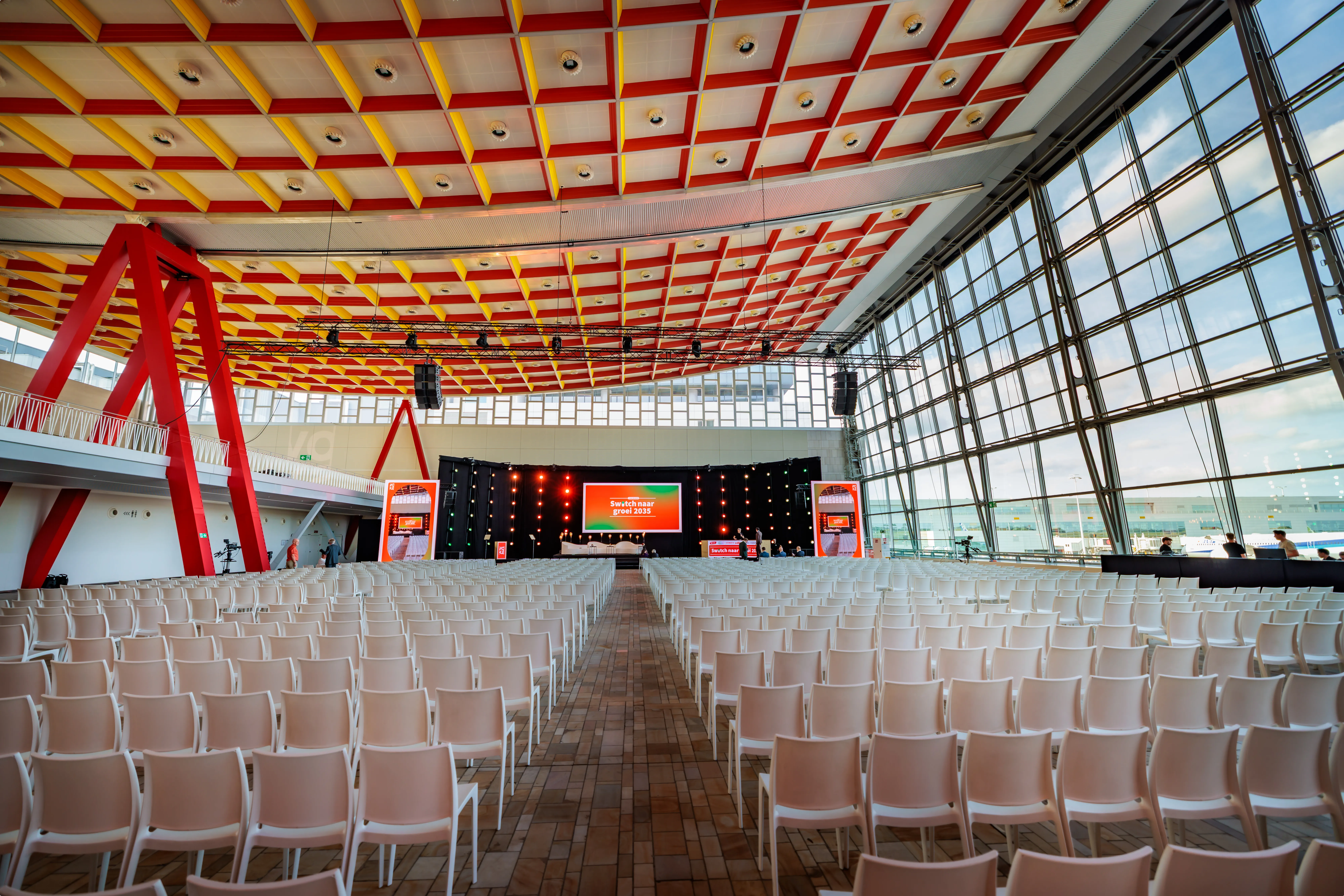 Spacious modern conference hall with rows of white chairs facing a stage and large windows on one side, featuring colorful ceiling patterns.