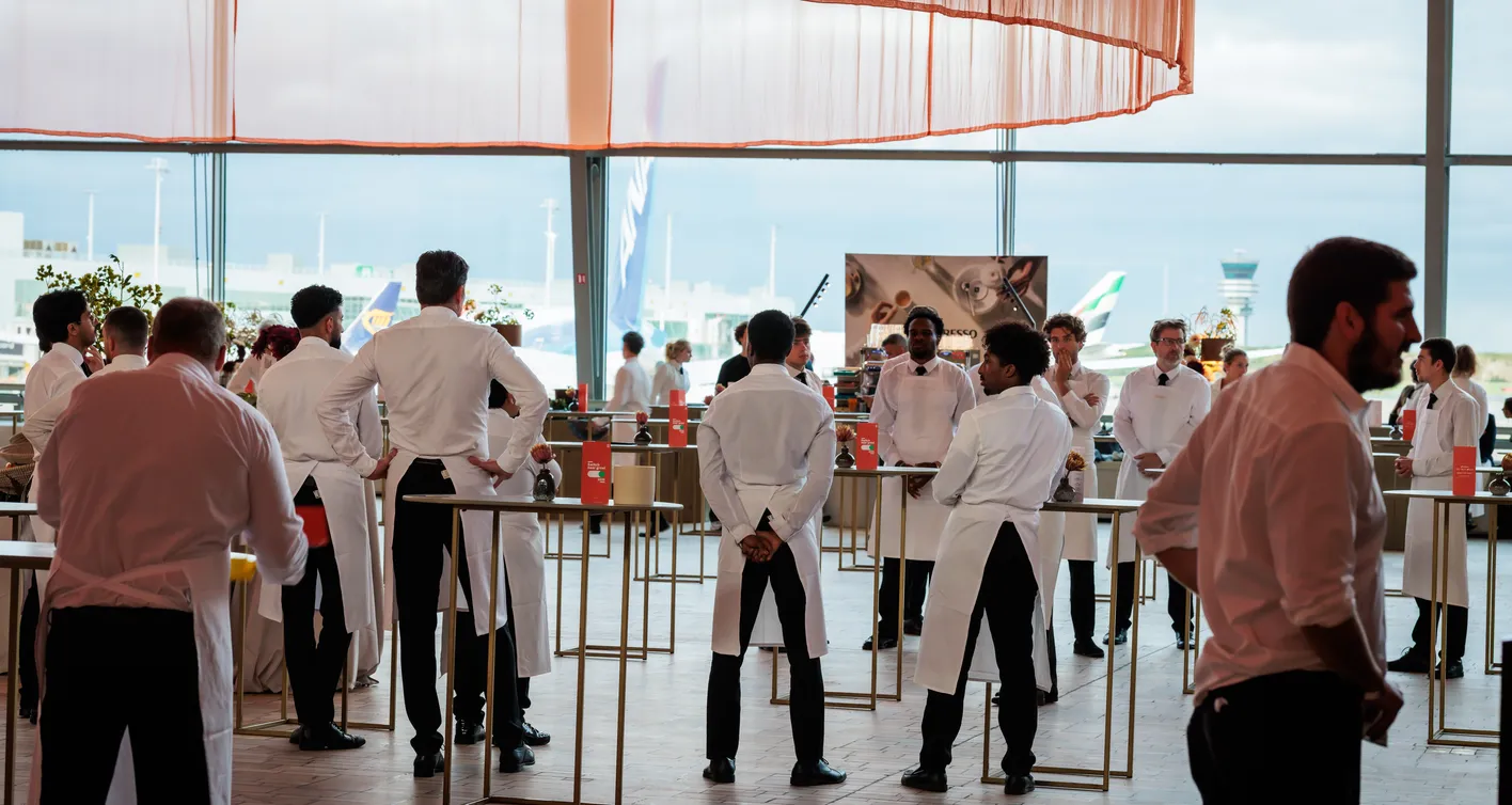 A group of servers in white shirts and aprons stand in a spacious, well-lit room, engaging in conversation and preparing for service.