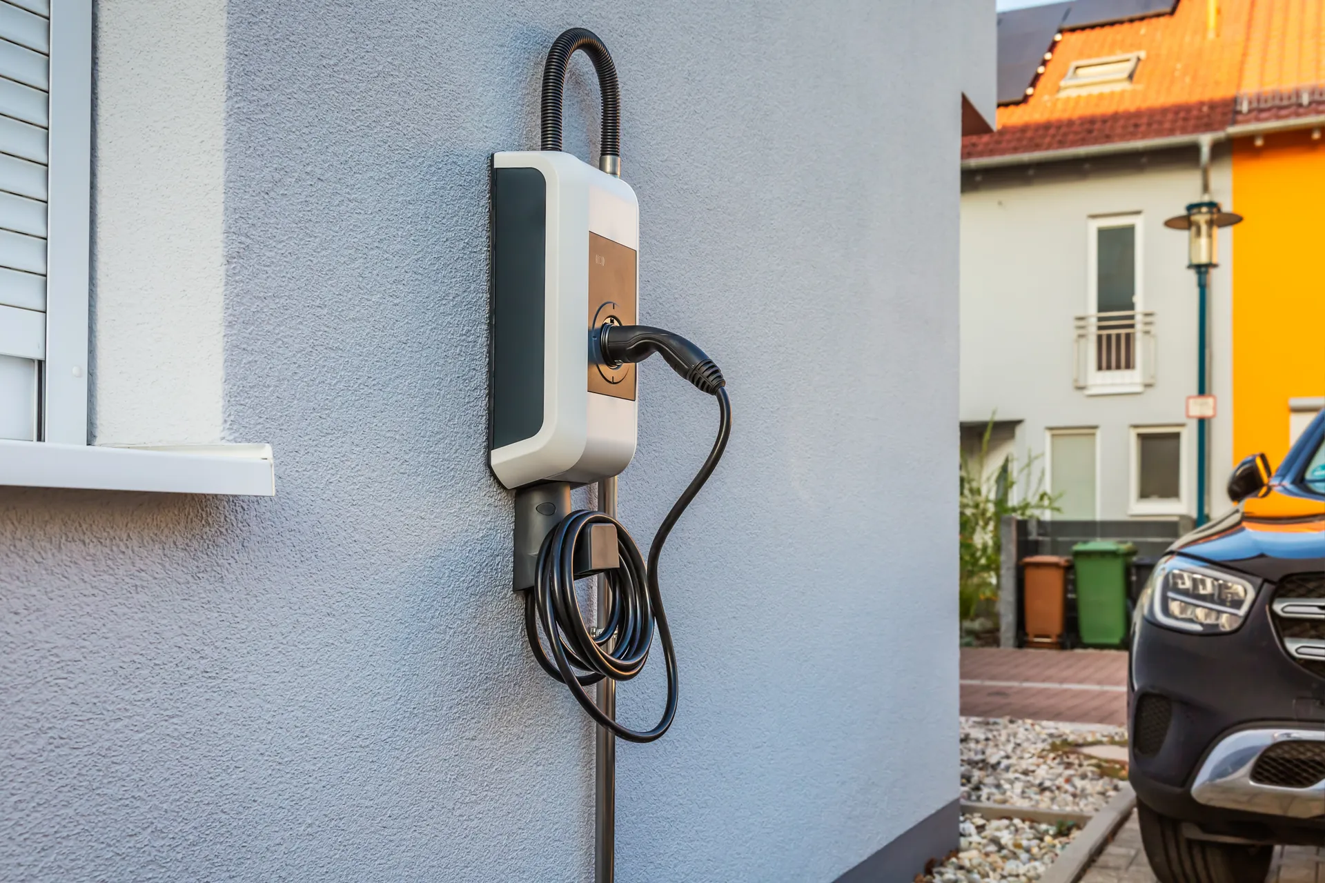 Eine an einer Hauswand montierte Ladestation für Elektrofahrzeuge mit einem Spiralkabel und einem Stecker. In der Nähe ist ein Auto teilweise zu sehen.