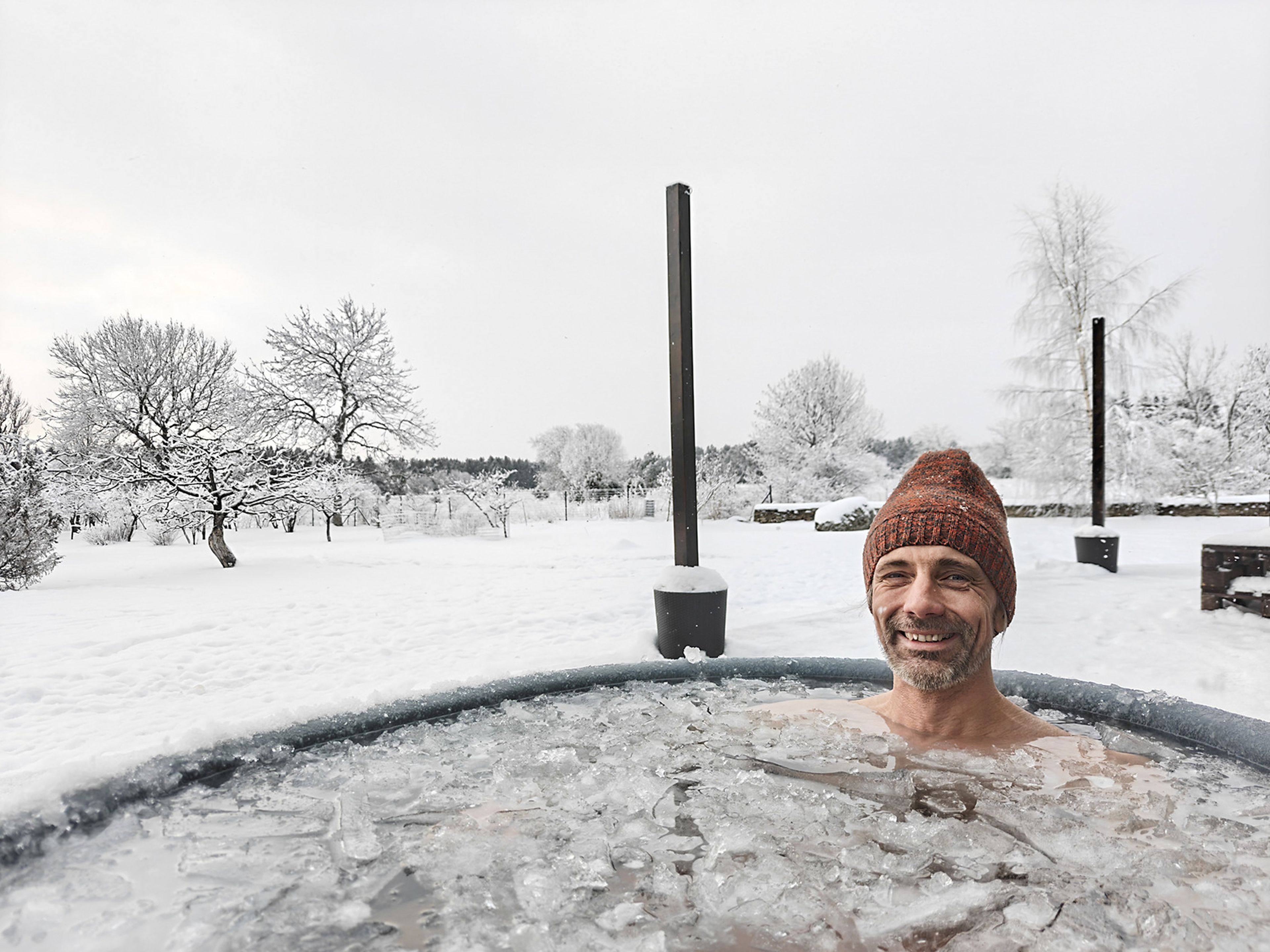 Ein Mann, der in einem eisigen Außenbad lächelt, umgeben von Schnee, mit einer braunen Strickmütze auf dem Kopf; im Hintergrund eine Winterlandschaft.