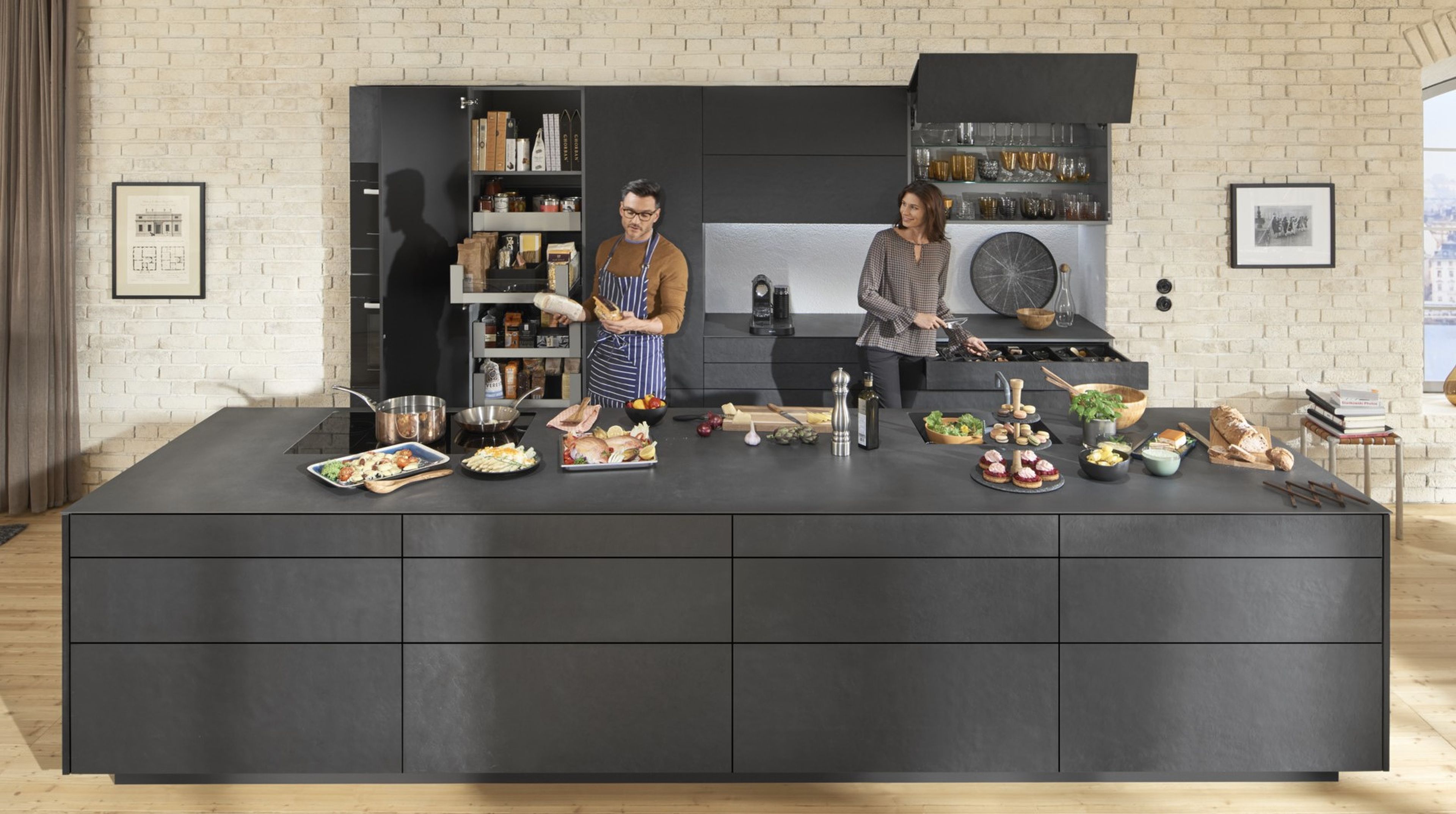 A man and a woman working together at an island kitchen with dark-coloured units.
