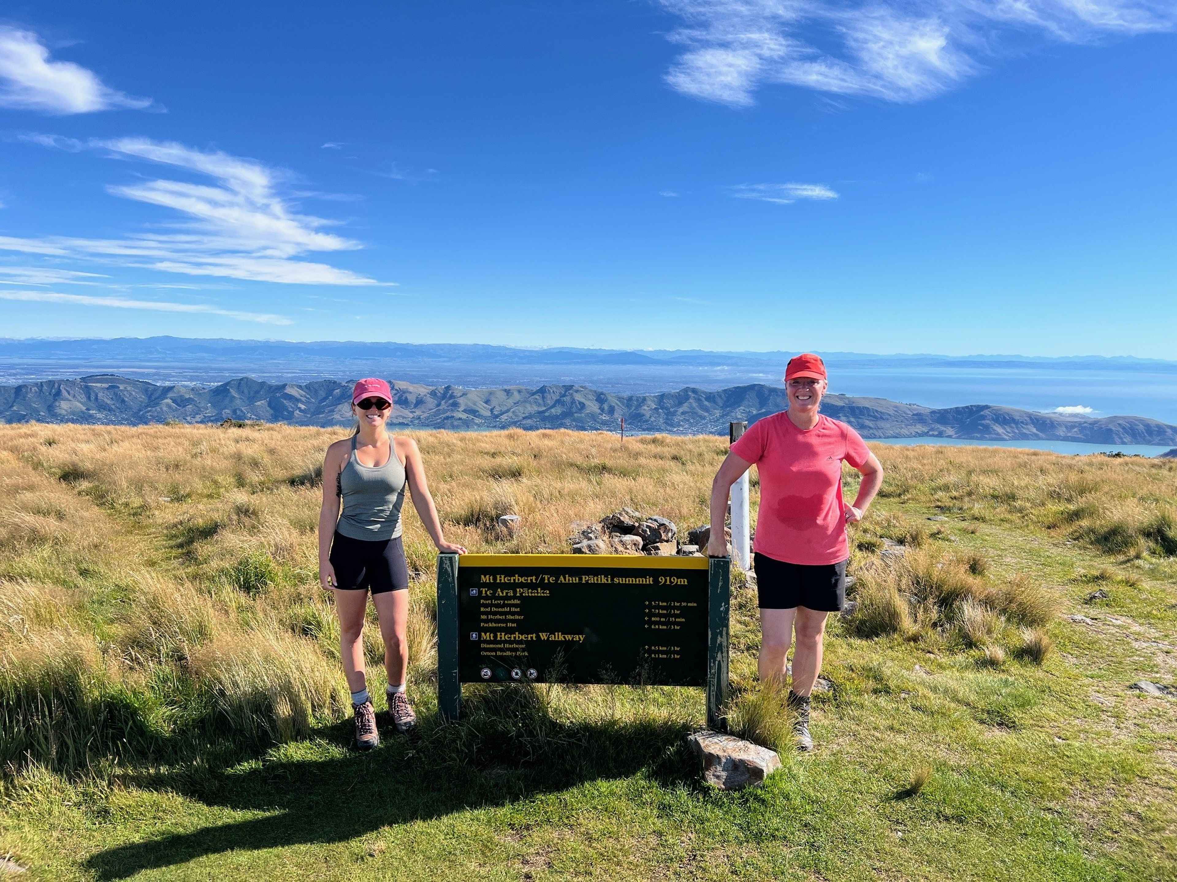 Two women in athletic wear stand beside a sign on a grassy hilltop, with a vast landscape and blue sky in the background.