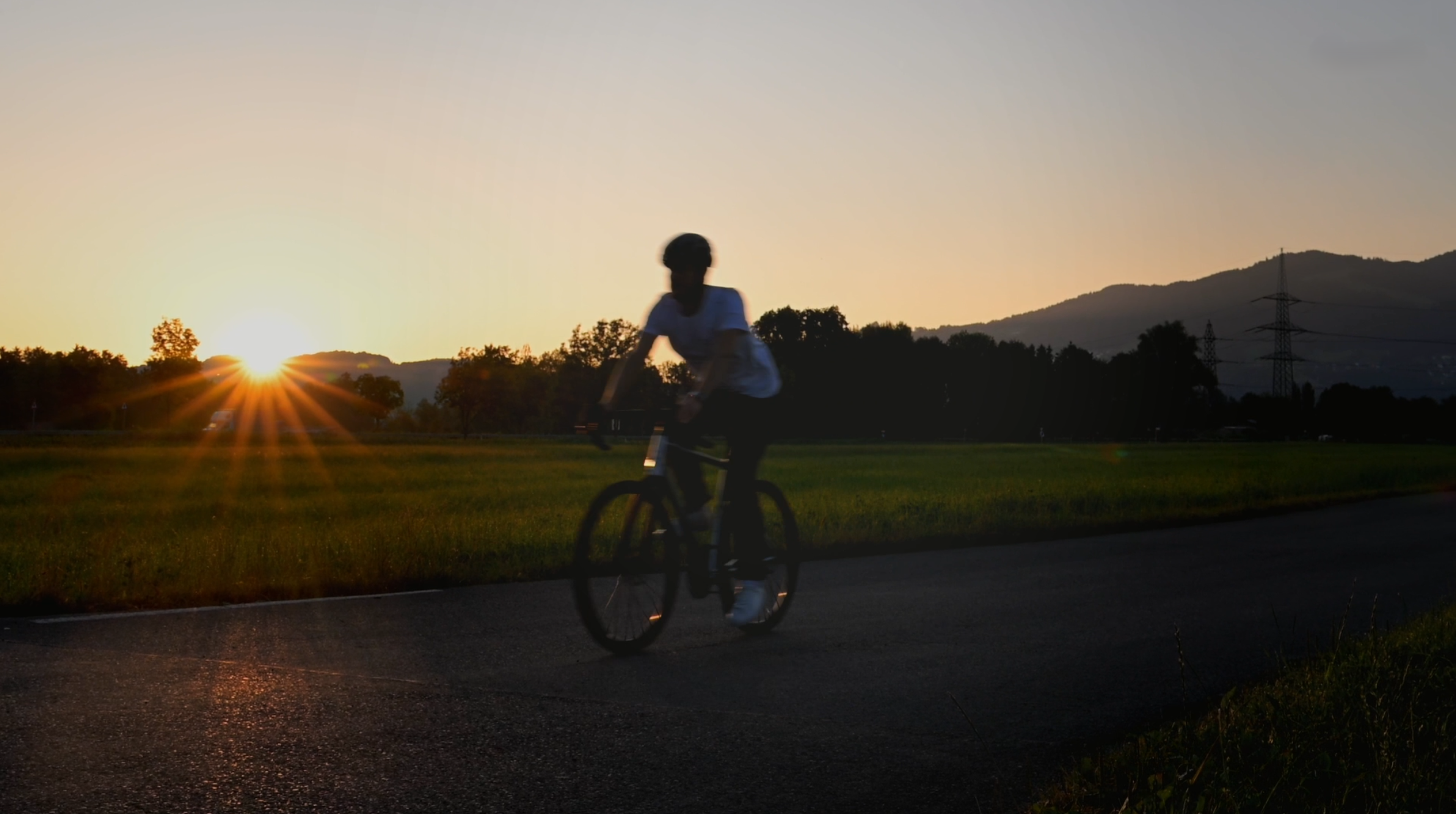 Ein Radfahrer auf einem Feldweg bei Sonnenuntergang, umgeben von Feldern und sich abzeichnenden Bäumen, mit Bergen im Hintergrund.