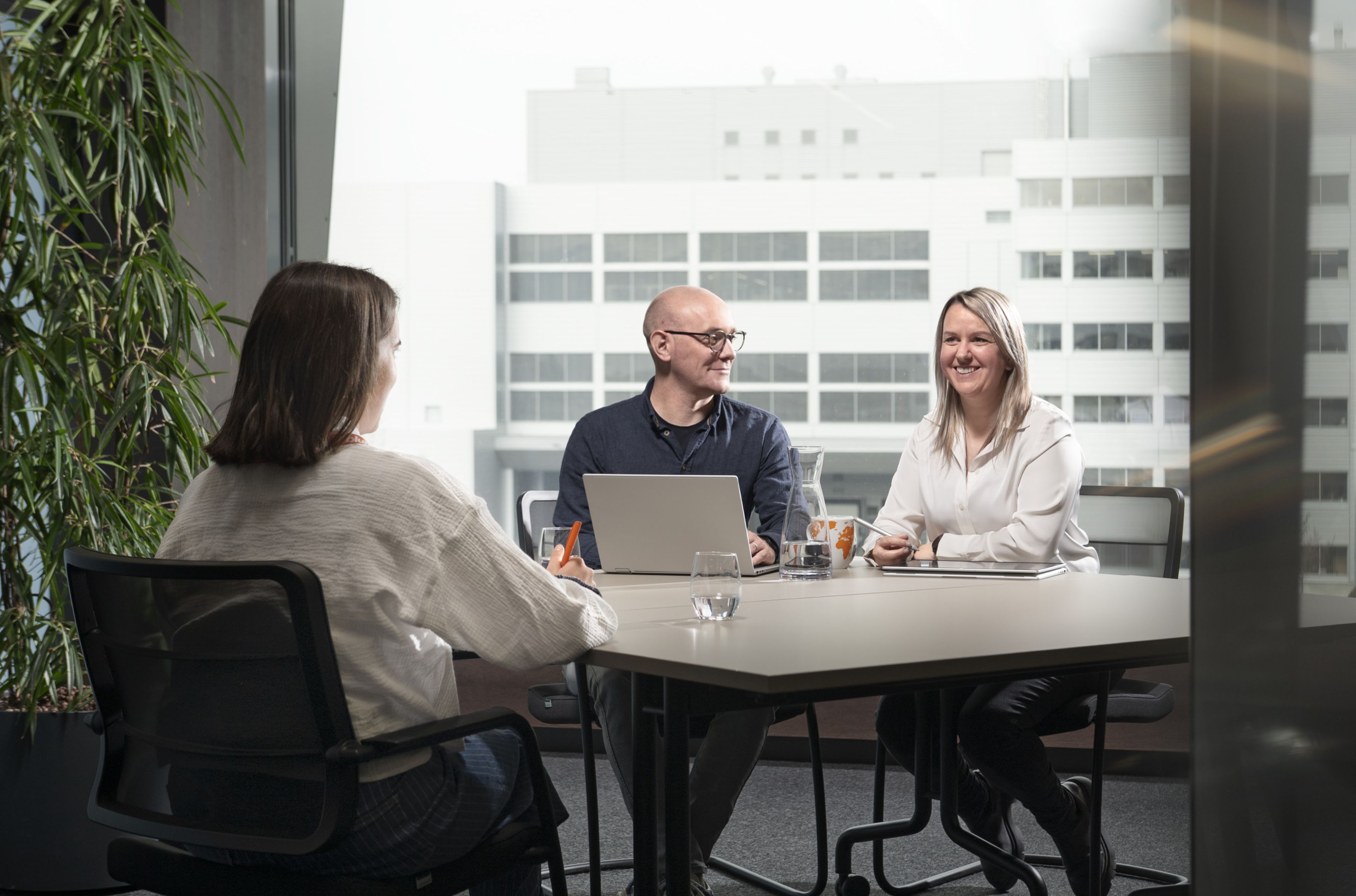 Drei Personen in einem modernen Büro, die eine Besprechung abhalten. Zwei sitzen mit ihren Laptops auf einer Seite des Tisches, während eine ihnen gegenüber sitzt.