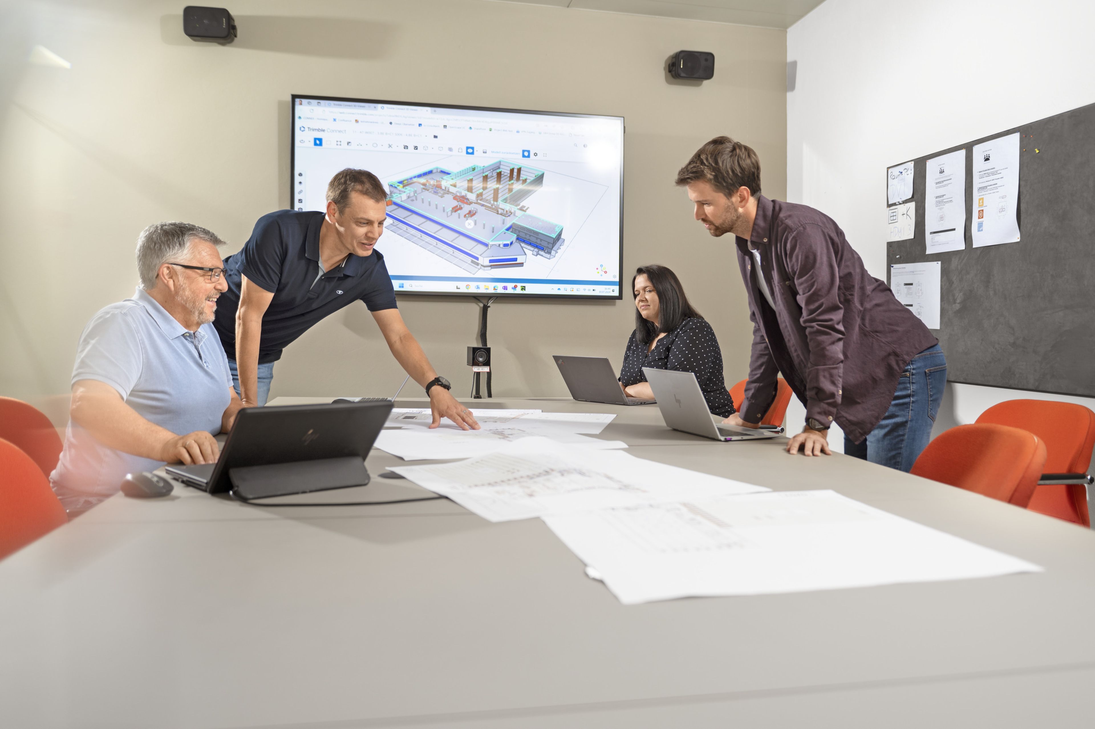 Four people are intently studying a building plan at a conference table. A 3D model of a building is visible in the background.