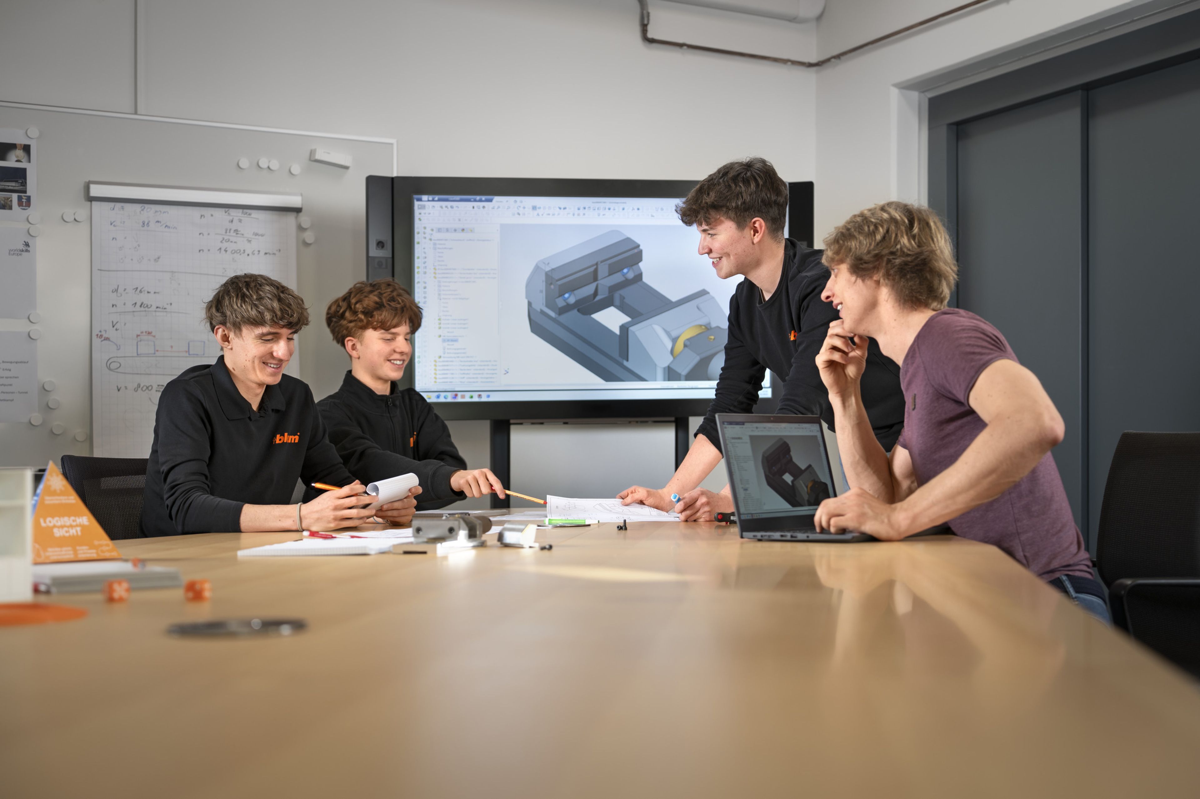 Abstimmung in der Konstruktionstechnik: Lehrlinge und Ausbilder besprechen gemeinsam ein technisches Bauteil und prüfen CAD‑Modelle sowie Zeichnungen in einem Besprechungsraum.