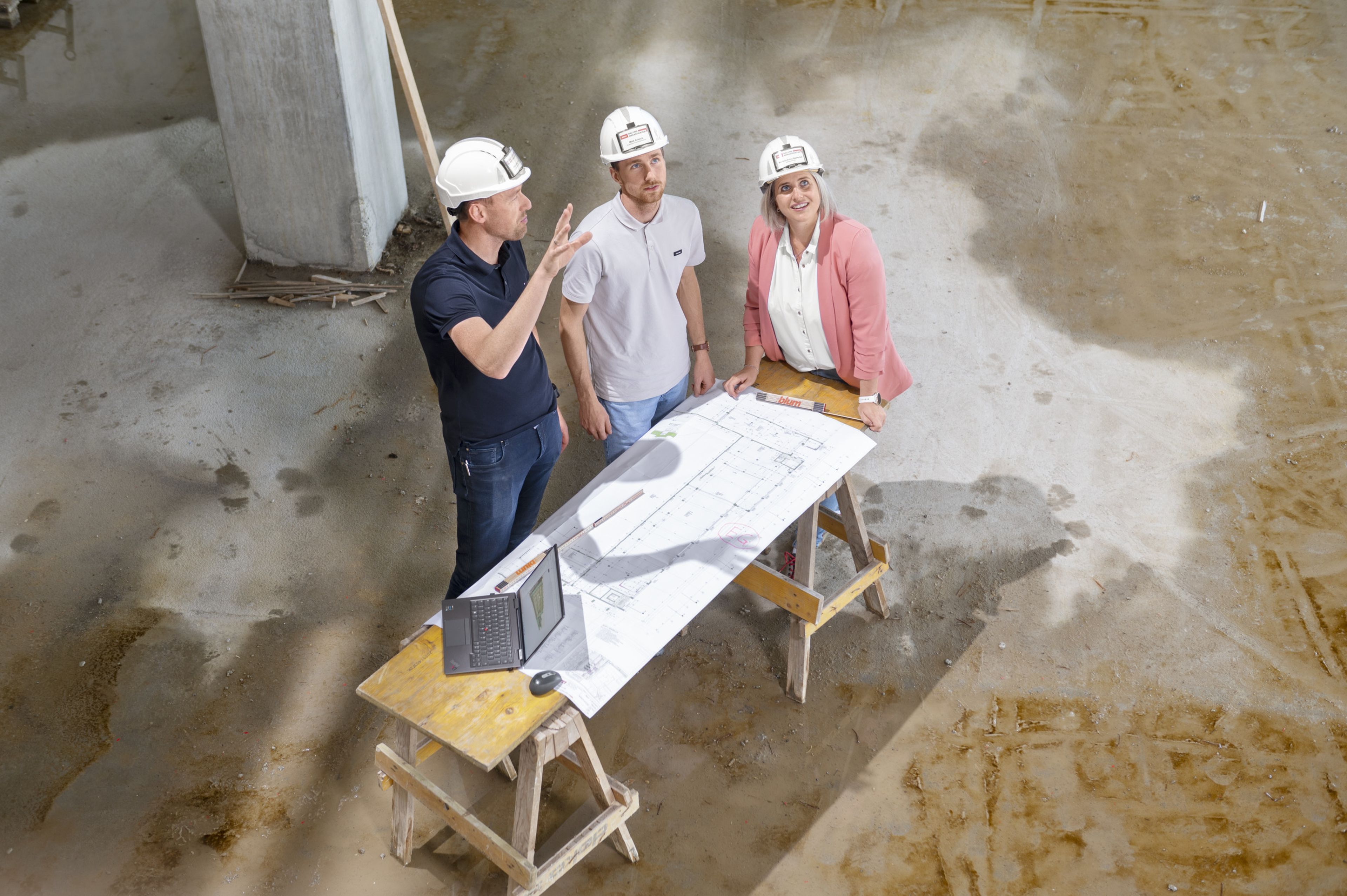Two men and a woman are standing in a construction site discussing the situation. In front of them, a building plan and a laptop are on a table.