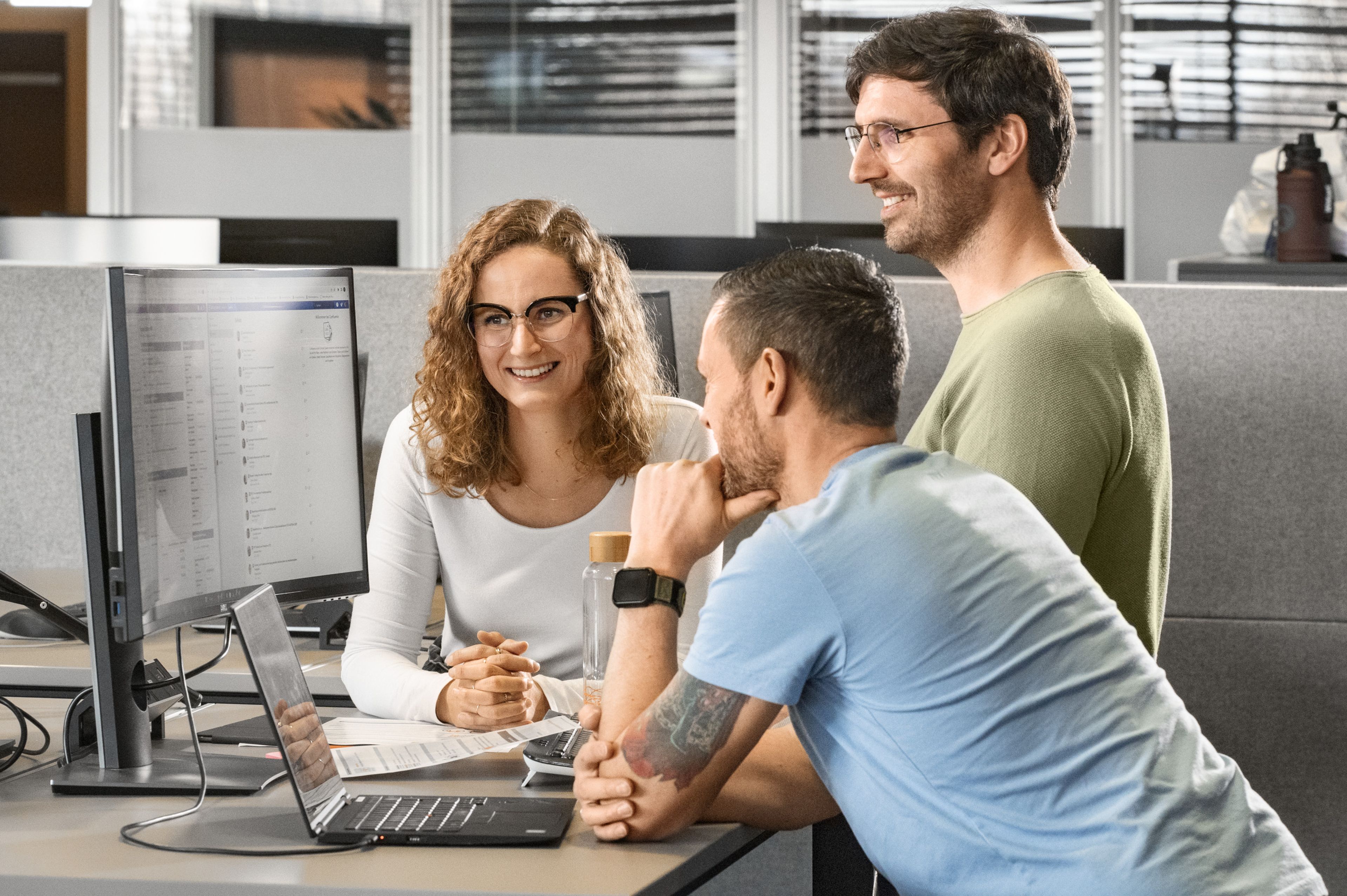 Eine Frau und zwei Männer schauen gemeinsam auf einen PC Bildschirm und lächeln