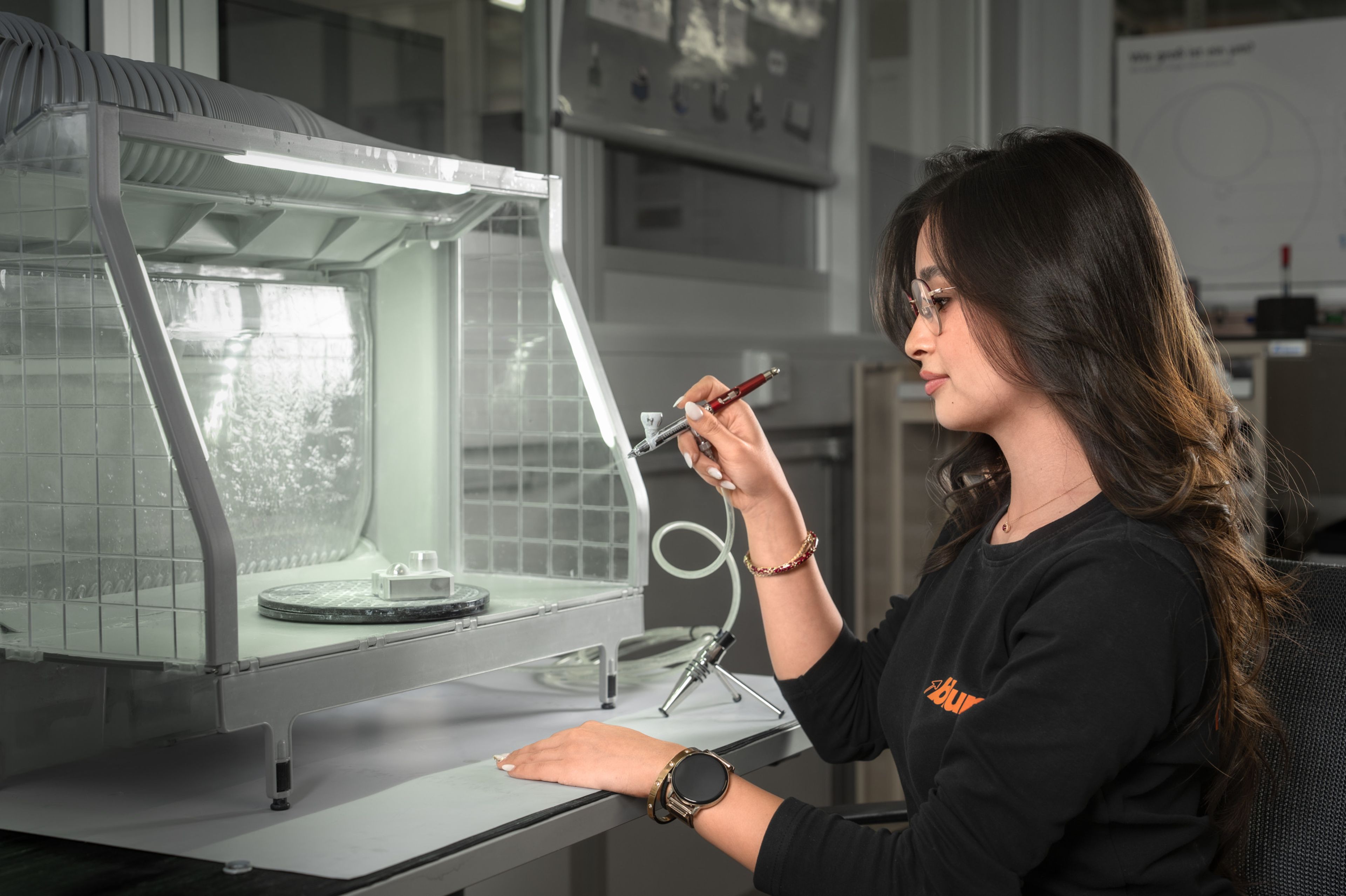 Auszubildende in der Ausbildung bei Blum im Beruf Fertigungsmesstechnik besprühen ein Bauteil mit einer Airbrush in einer Messkabine, um die Oberfläche für die anschließende digitale Vermessung vorzubereiten.