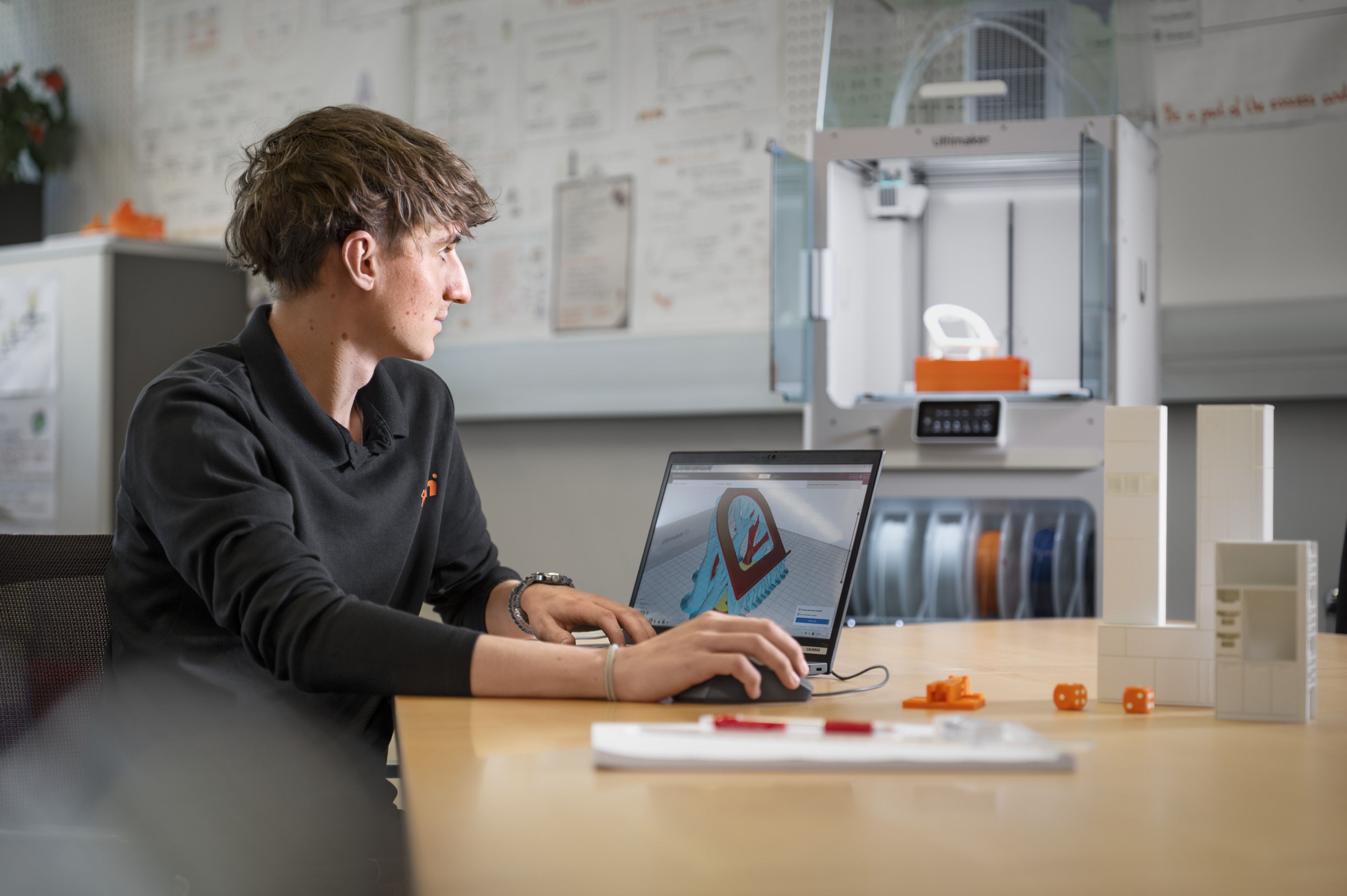 Lehrling in der Konstruktionstechnik arbeitet am Laptop an einem 3D‑Modell, während im Hintergrund ein 3D‑Drucker Prototypenbauteile für die technische Entwicklung erstellt.