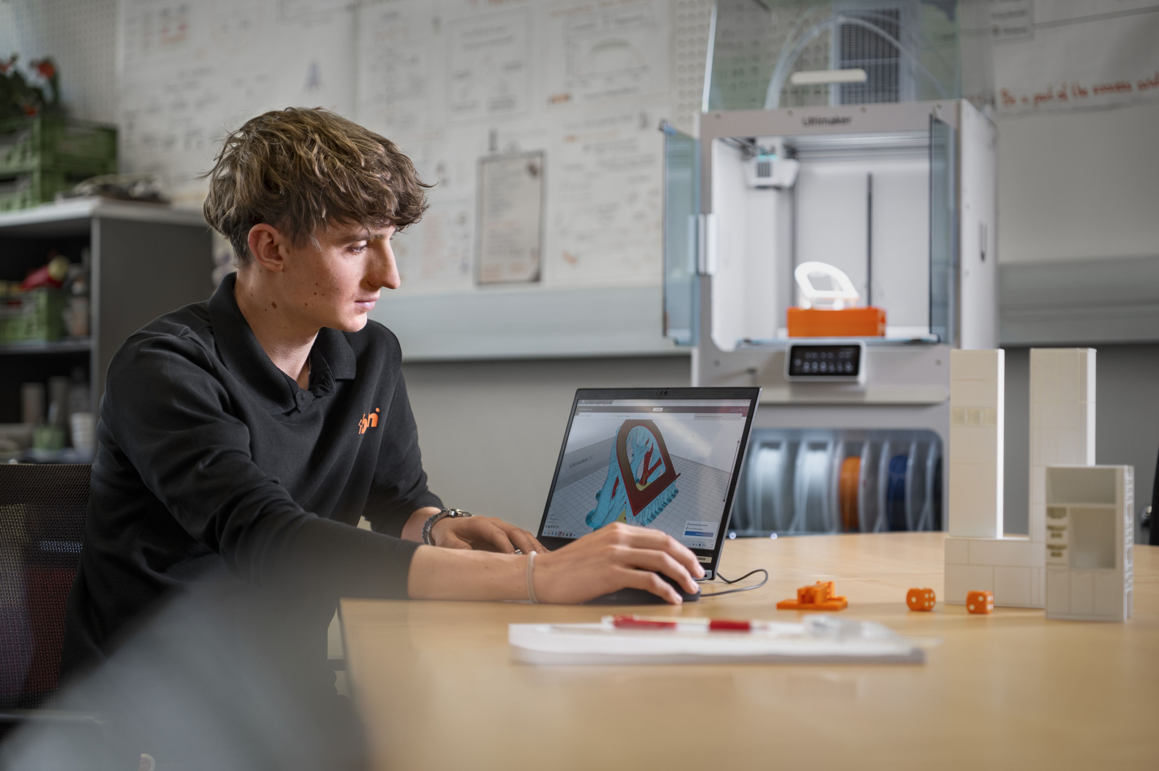 Trainees der Dualen Akademie Konstruktionstechnik arbeiten bei Blum an einem CAD‑Entwurf am Laptop. Auf dem Tisch stehen 3D‑gedruckte Bauteile und technische Modelle, im Hintergrund ein moderner Arbeits‑ und Entwicklungsbereich.