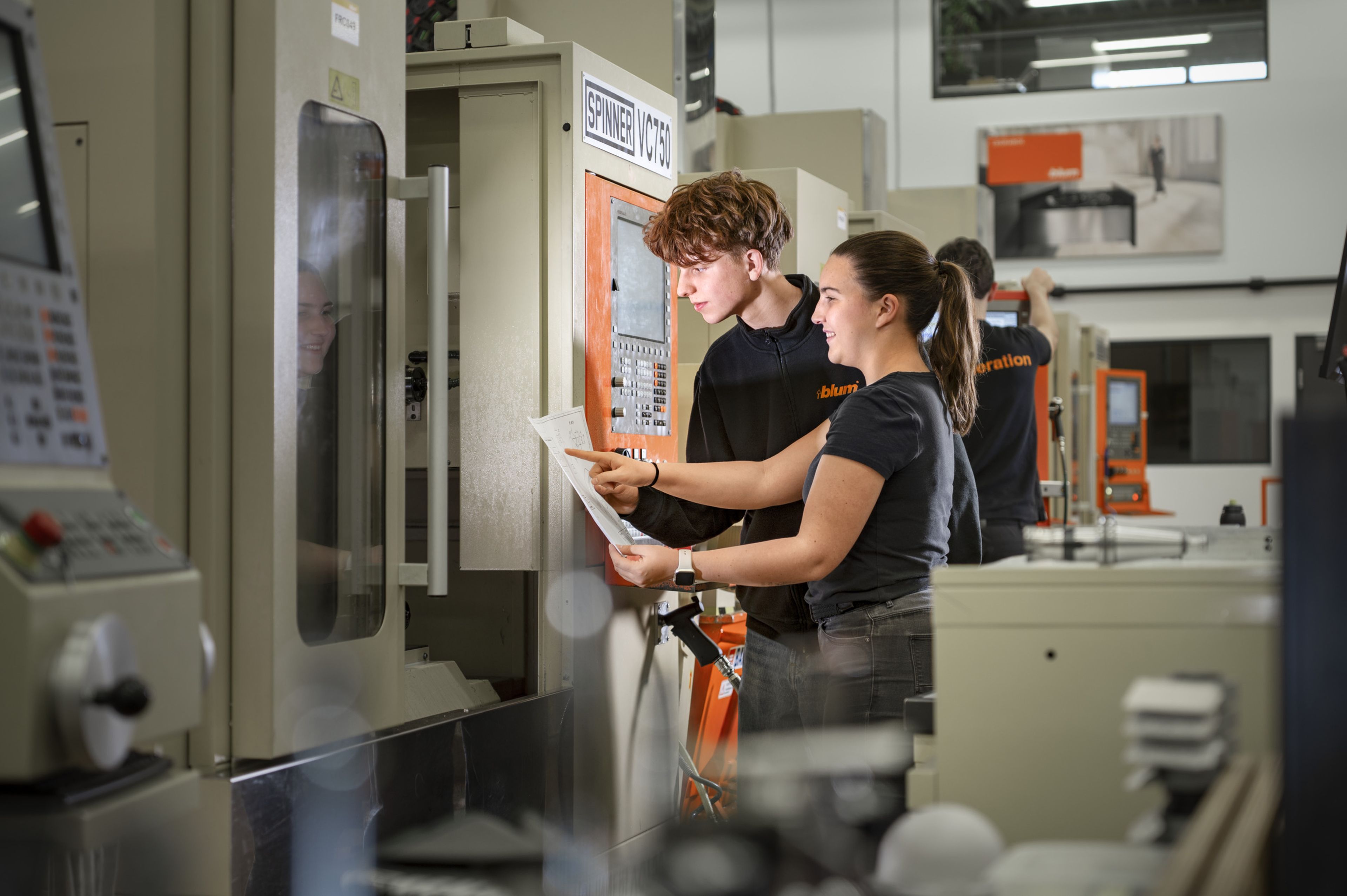 Konstruktionstechnik‑Lehrling und Auszubildende in der Zerspanung prüfen gemeinsam eine technische Zeichnung und die CNC‑Programmierung an einer Produktionsmaschine in der Fertigung.