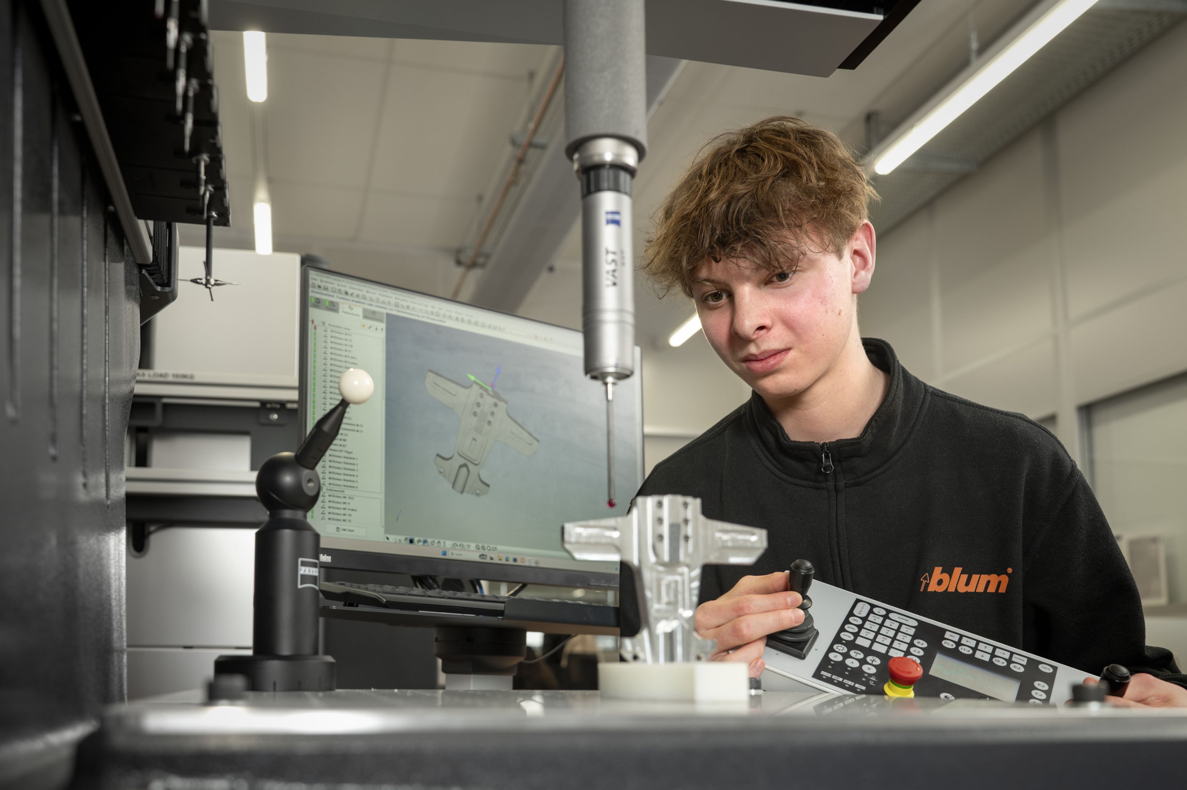 Auszubildende in der Ausbildung bei Blum im Beruf Fertigungsmesstechnik messen ein Präzisionsbauteil an einem Koordinatenmessgerät, steuern die Messung über das Bedienpanel und werten die Messergebnisse am Computer anhand eines CAD‑Modells aus.