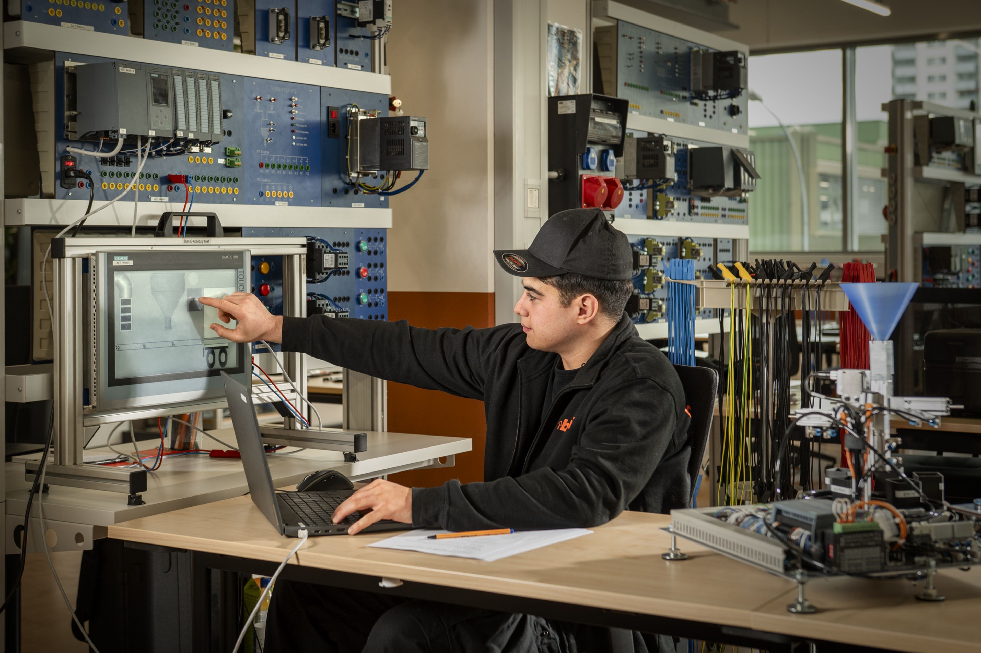 Auszubildender in der Steuerungstechnik bei Blum programmiert und testet an einem Industriearbeitsplatz eine speicherprogrammierbare Steuerung über einen Touchscreen, umgeben von Schaltschränken, Verdrahtungen und Steuerungskomponenten in der Ausbildungswerkstatt.