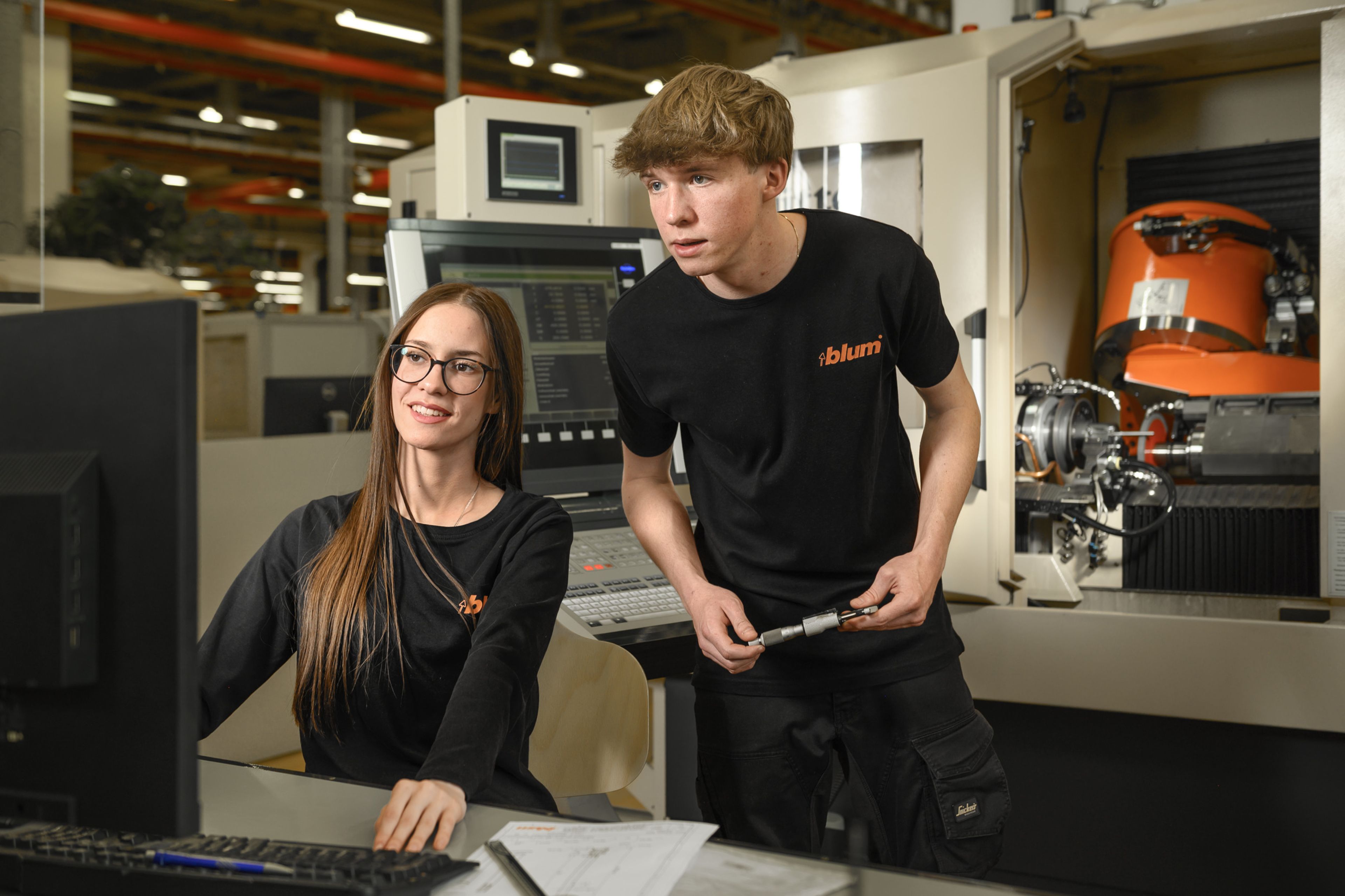 Zwei Auszubildende in der Zerspanungstechnik bei Blum planen und überwachen einen Schleifprozess an einer CNC-Schleifmaschine. Das Bild zeigt die Zusammenarbeit bei der Präzisionsbearbeitung von Metallbauteilen, die digitale Maschinensteuerung und die praxisnahe Ausbildung in der Zerspanungstechnik bei Blum.