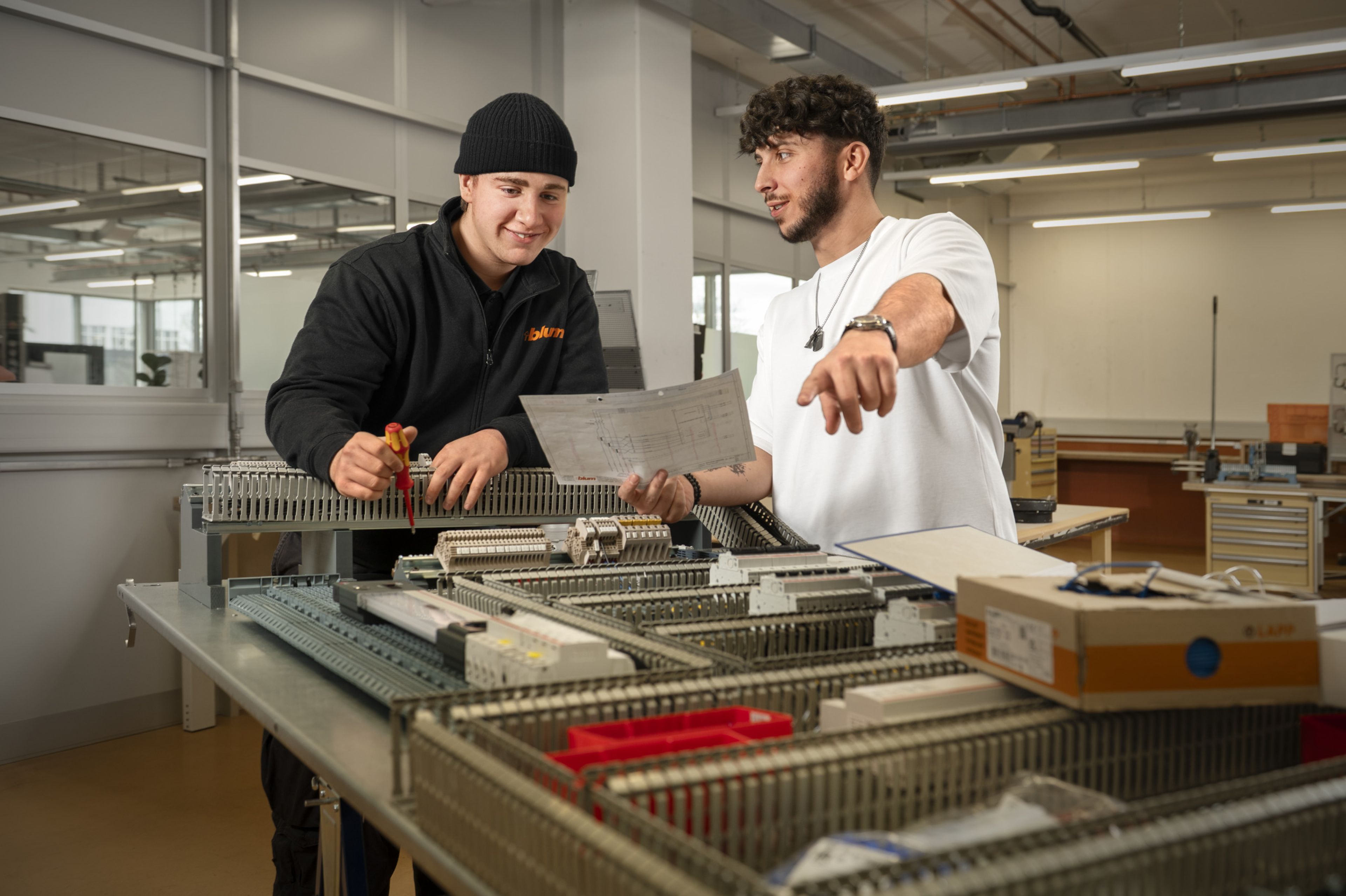 Ein Auszubildender und ein Ausbilder bei Blum montieren und verdrahten gemeinsam einen offenen Schaltschrank auf einem Arbeitstisch und prüfen anhand eines Schaltplans die Anordnung von Klemmen, Leitungen und elektrischen Komponenten in der Ausbildungswerkstatt.