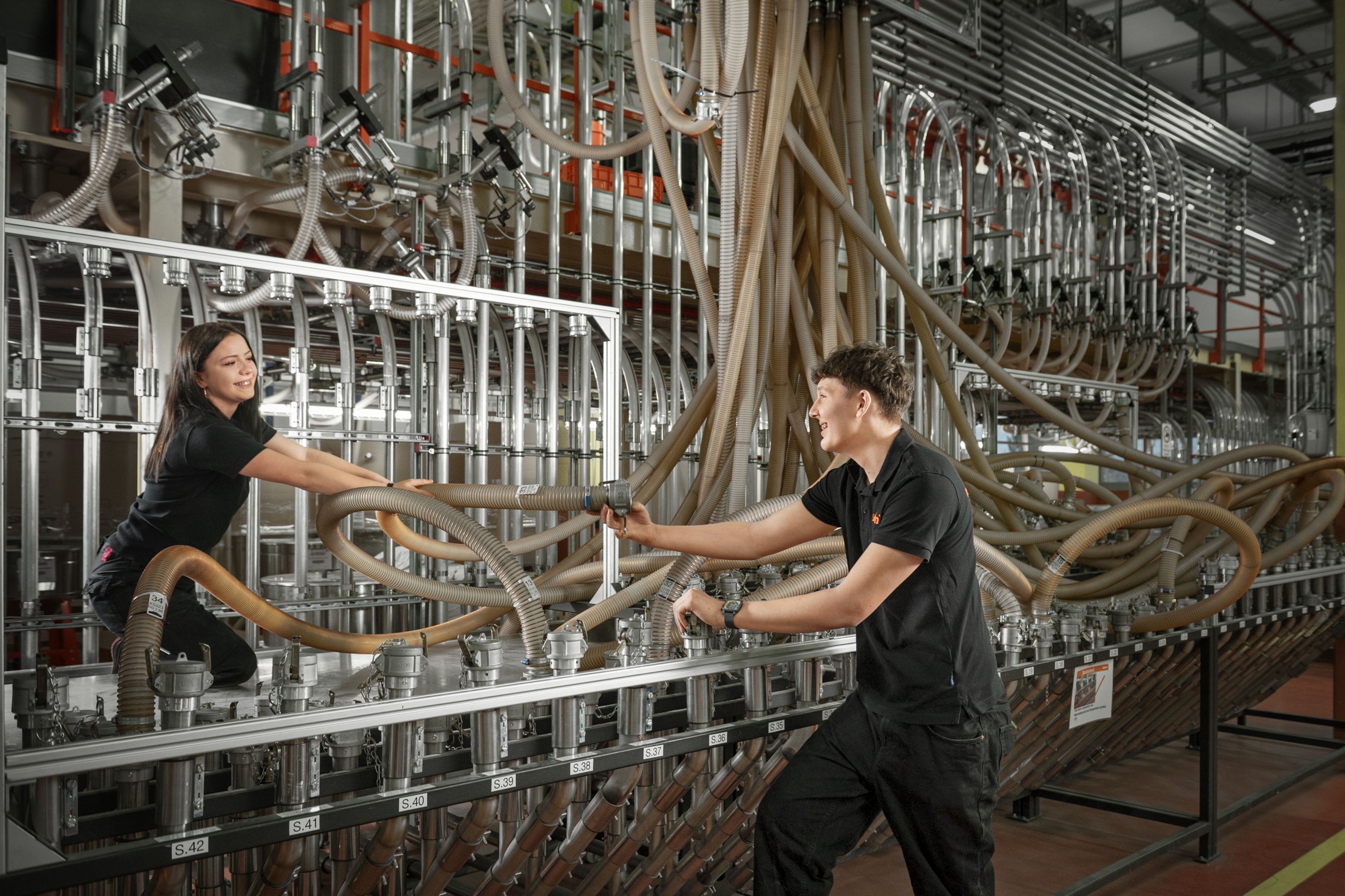 Das Foto zeigt zwei Auszubildende die gerade die Leitungen des Kunststoffgranulats neu anstecken bzw. nachfüllen. 
