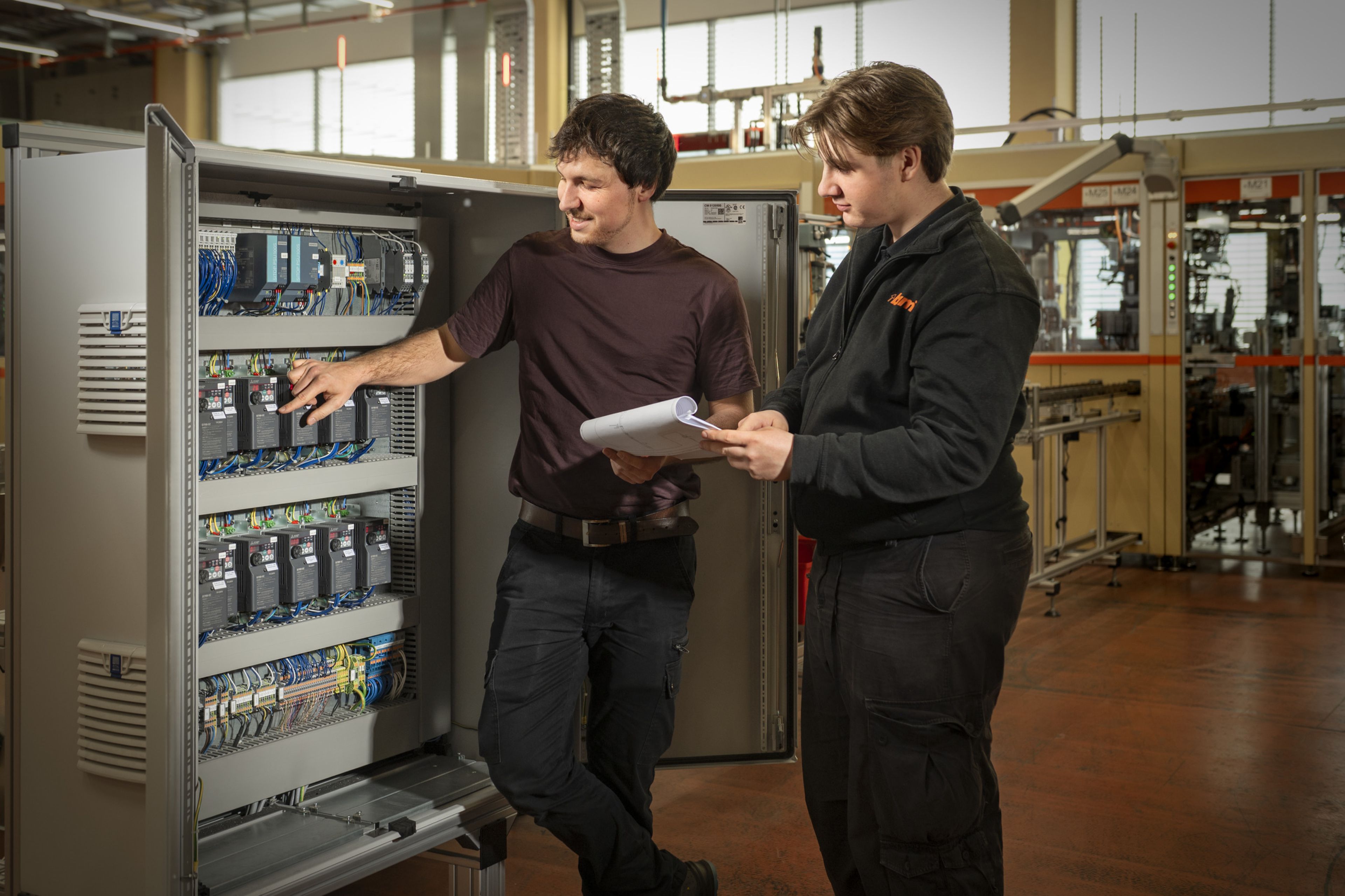 Ein geöffneter Schaltschrank ist in einer industriellen Montageumgebung installiert, während zwei Personen die verbauten Steuerungs- und Antriebskomponenten prüfen und anhand von Unterlagen Einstellungen und Funktionen der Anlage kontrollieren.